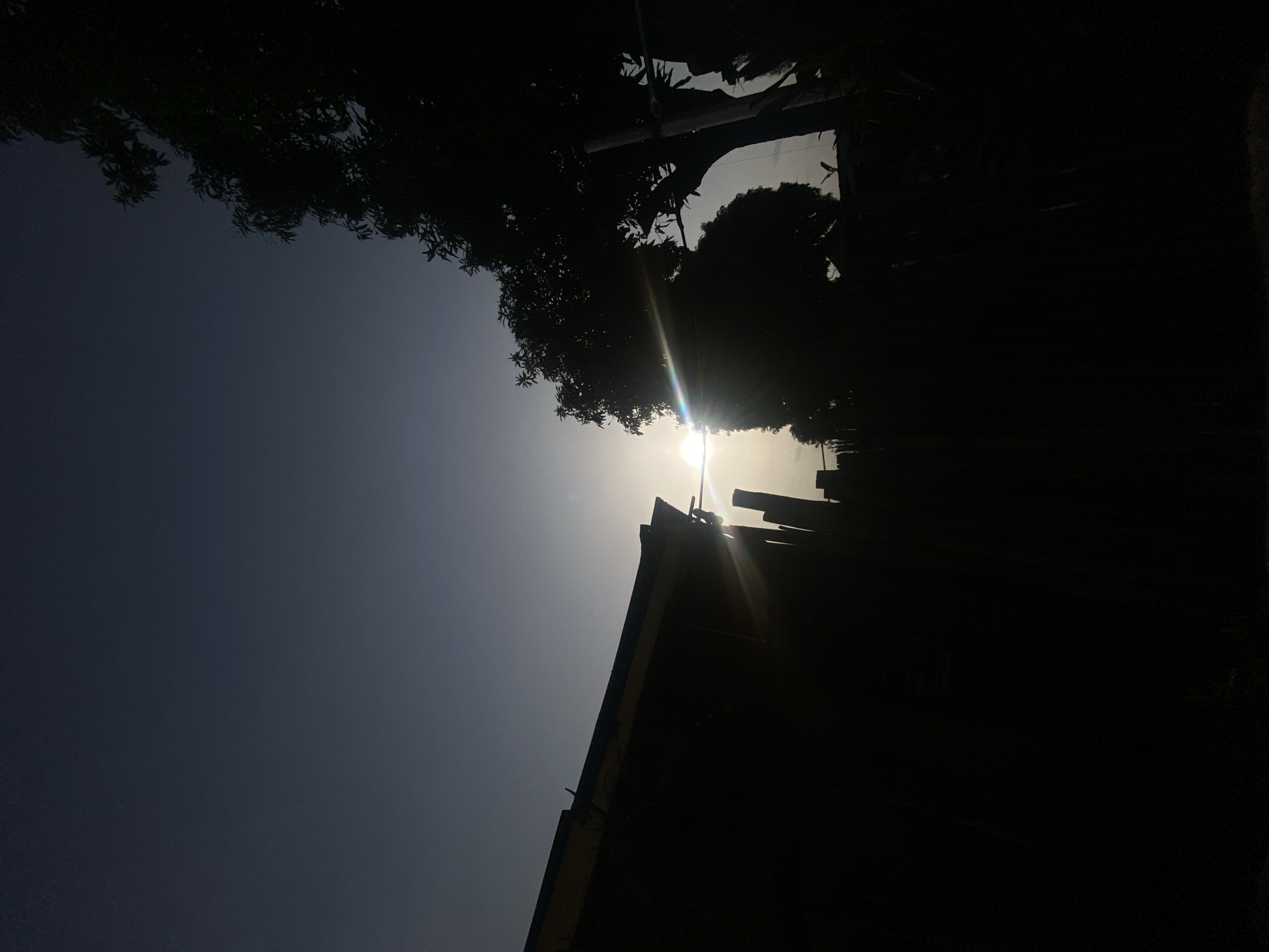 The image, taken outdoors, depicts a low-angle shot of a bright sun shining through trees and the edge of a building. The building and trees appear as dark silhouettes against the bright sky. The location is assumed to be in Barden Yana, Nigeria. There are no people visible.
