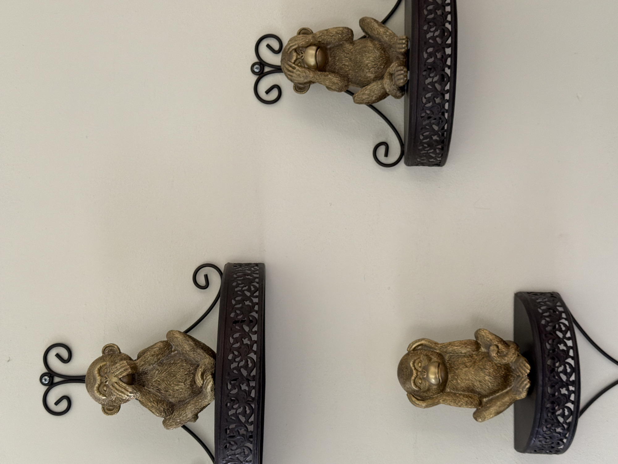 The image shows three decorative monkey figurines mounted on a beige wall. Each figurine is a metallic golden color and depicts a different pose from the "See no evil, hear no evil, speak no evil" proverb. The monkey on the upper right is covering its eyes, the one on the lower left is covering its mouth, and the one on the lower right is covering its ears. Each monkey is perched on a small, semicircular shelf with a black, ornate metal backing and scrolled wrought iron supports. There is no evidence in this image indicating the location of Cuautla, Mexico.