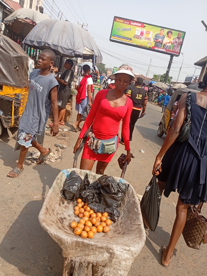 The image depicts a bustling street scene in Onitsha, Nigeria, under direct sunlight, indicated by sharp shadows. In the immediate foreground, a metal wheelbarrow, lined with a tan sack, contains numerous small, round, orange-yellow fruits and several black plastic bags.
A woman, positioned centrally, pushes the wheelbarrow. She wears a long-sleeved red shirt, a short red skirt, a light-colored visor, and a multi-colored fanny pack around her waist. She looks directly towards the viewer. To her left, a young male, possibly a teenager, walks barefoot, wearing a grey sleeveless shirt with "CAROLINA H*COCKS" text and torn blue denim shorts. He gazes upwards and to his left.
On the right side of the frame, a second woman walks away from the viewer, wearing a dark blue dress and light brown sandals. She carries a black plastic bag in her right hand and a brown, patterned handbag on her left shoulder.
The background reveals a busy street with additional pedestrians, including a man in a black and yellow jersey with the number "12" on the back, walking away. Several makeshift market stalls or shelters with grey tarpaulin canopies are visible. A yellow and black tricycle (keke napep) is partially visible on both the left and right sides of the street. Overhead power lines crisscross the sky. In the mid-distance, a large billboard advertises "Coafal SoftGel" with images of people and product, displaying the slogan "No Tremor!". The road surface appears worn asphalt with scattered debris.