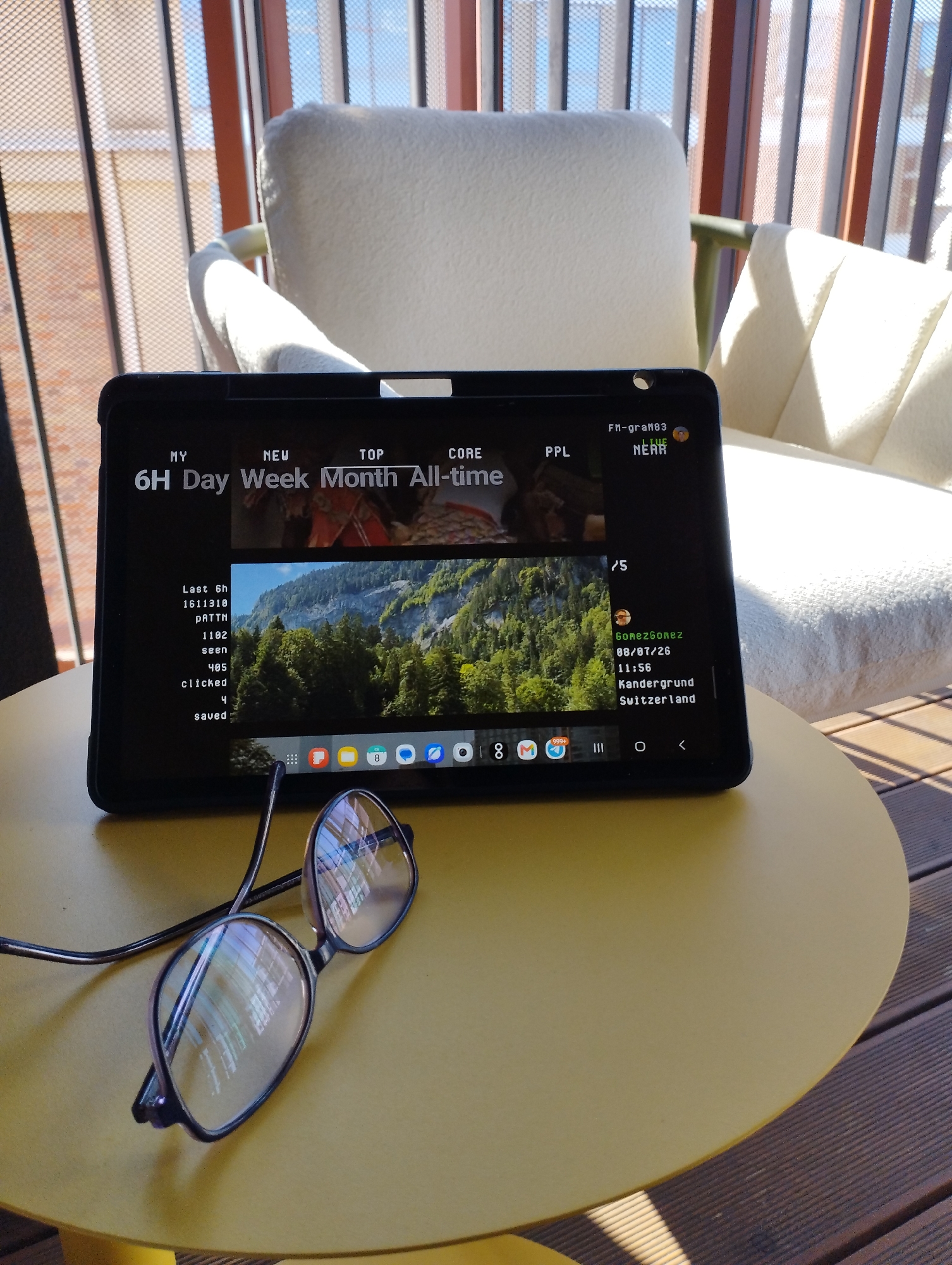 In the foreground, a pair of eyeglasses with dark frames and clear lenses rests on a yellow, circular table. The lenses of the eyeglasses reflect the scene, showing streaks of light and some indistinct objects. The table is positioned on a wooden deck. In the background, a black tablet is propped up, displaying a screen with text and an image of a mountainous landscape with lush green trees and a clear blue sky. The tablet screen shows menu options like "MY," "NEW," "TOP," "CORE," and "PPL," along with time-based filters such as "6H," "Day," "Week," "Month," and "All-time." Below these, there is data indicating "Last 6h," "1611310," "pATTN," "1102," "seen," "405," "clicked," and "saved." Further down the screen, there is information that appears to be a user profile with a name, date, time, and location: "GomezGomez," "08/07/26," "11:56," "Kandergrund," and "Switzerland." There are also icons for various applications at the bottom of the tablet screen, including what looks like a web browser, email, and calendar. The tablet is positioned in front of a cream-colored armchair, and behind the armchair, there is a structure with vertical elements and some sunlight filtering through, suggesting an outdoor or semi-outdoor setting. The overall scene is bright and appears to be daytime.