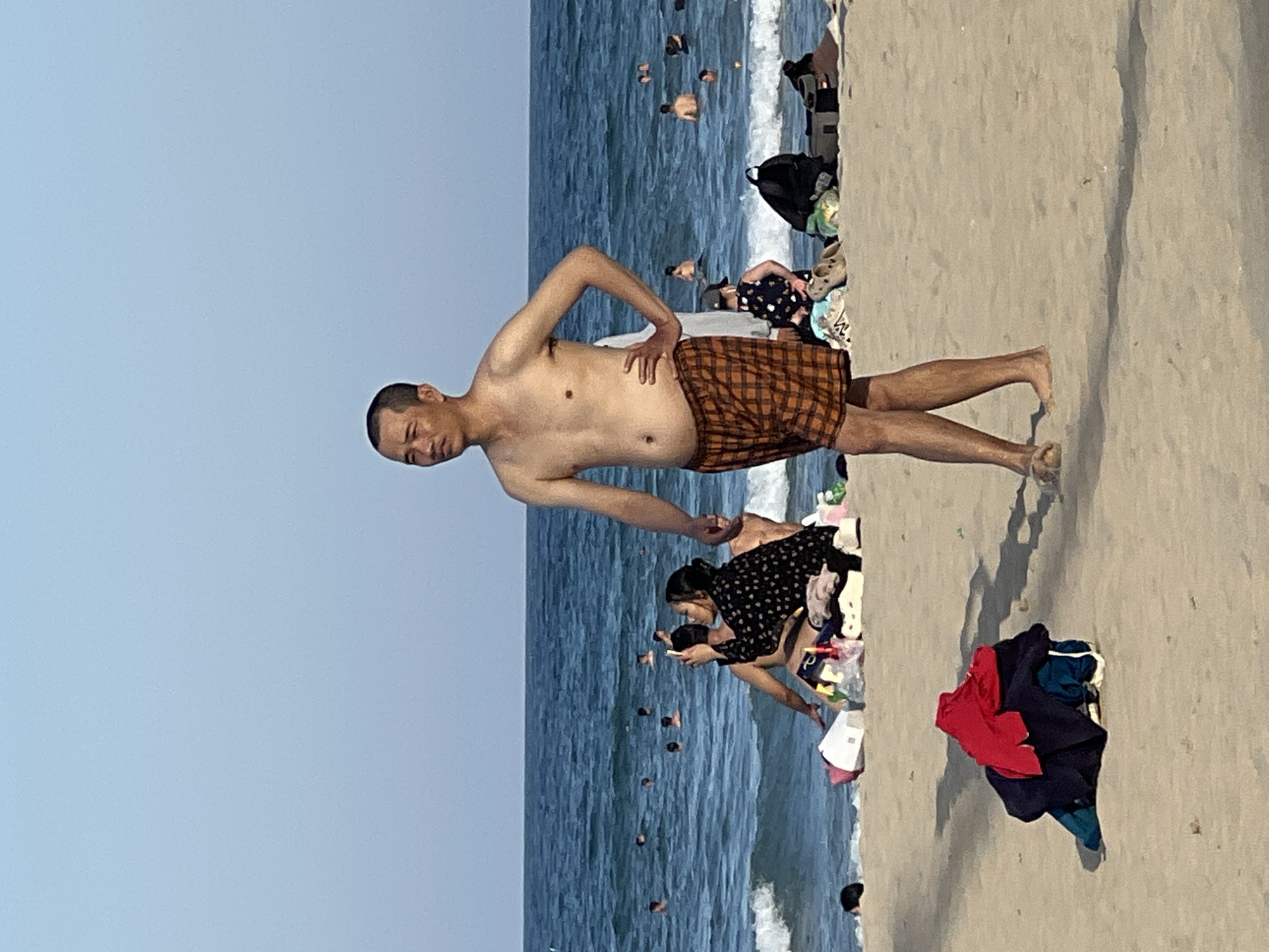 The image depicts a beach scene in Đà Nẵng, Vietnam. A shirtless man wearing plaid shorts stands on the sand, facing to his left, with one hand on his hip. In the background, the ocean is populated with numerous swimmers. Further ashore, a group of people are seated on the sand, with a woman in a black patterned dress holding up a mobile phone. Several bags and items of clothing are scattered on the sand in front of them. In the foreground, a pile of folded clothes, including a red shirt, lies on the sand. The sky is clear and blue, indicating a sunny day.