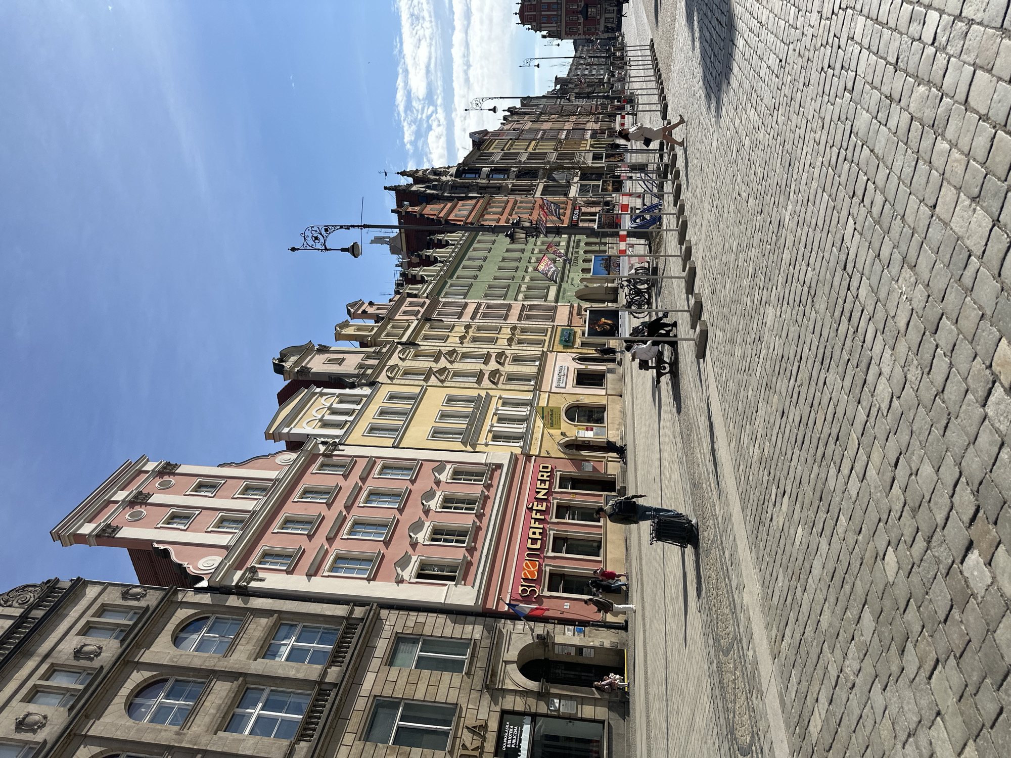 The image depicts a street scene in Wrocław, Poland, showcasing the colorful facades of buildings lining the Rynek (Market Square). The buildings display a variety of architectural styles and pastel hues, including pink, yellow, green, and brick red. The street is paved with cobblestones and is populated by pedestrians. A "Green Caffè Nero" sign is visible on one of the buildings. Metal barriers and bicycle racks line the edge of the pedestrian area. Street lamps are present along the street. The sky is blue with some clouds visible in the distance.