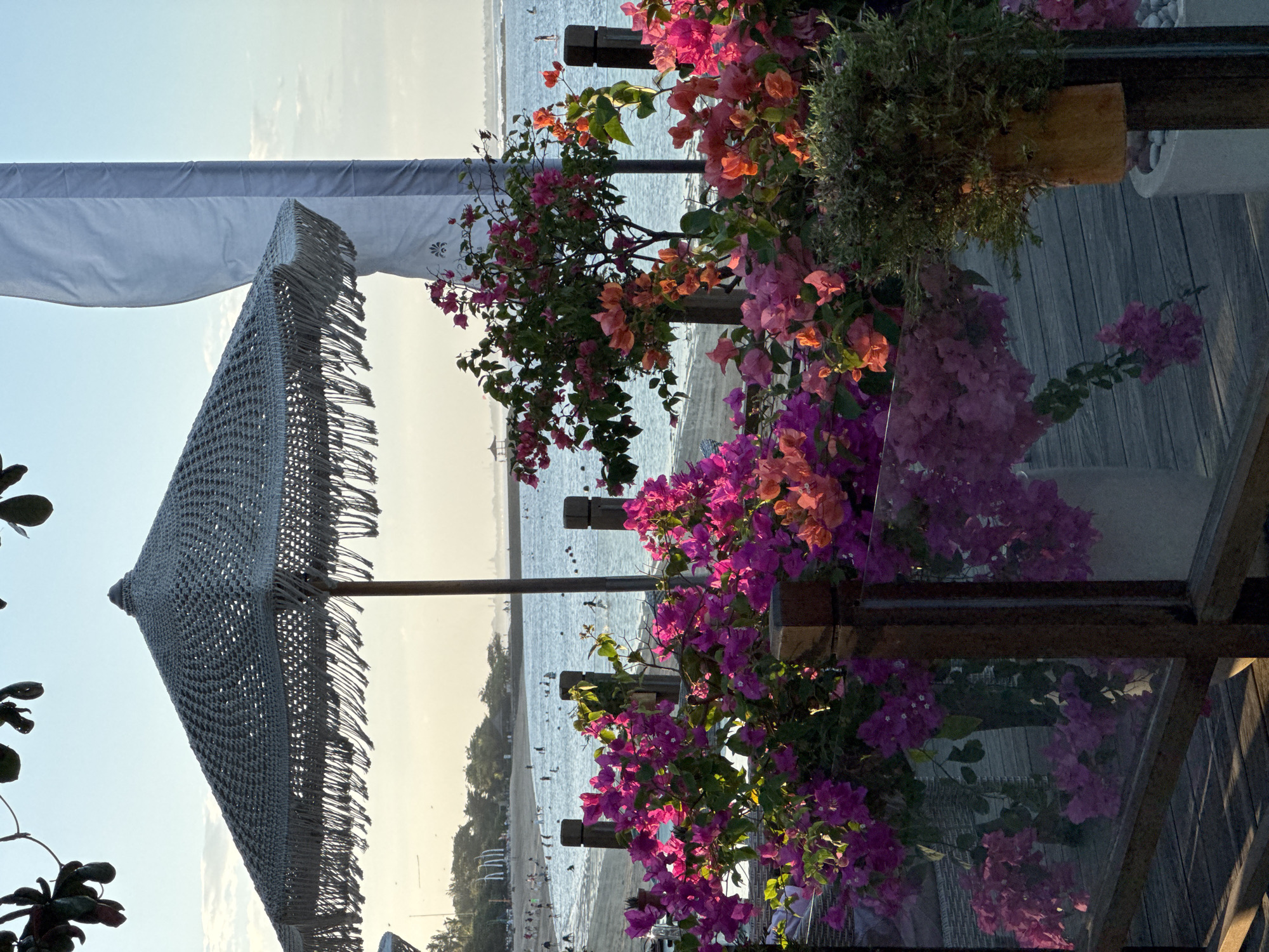 The image shows a view from a wooden deck overlooking Kuta Beach in Kuta Selatan, Indonesia. In the foreground, vibrant pink and orange bougainvillea flowers are in bloom, with some spilling over a glass railing and onto the wooden planks. Behind the flowers, a large thatched umbrella with fringe details is partially visible. Further back, the ocean meets a sandy beach with numerous people swimming and relaxing. The sky is a soft blue with scattered clouds, and the sun appears to be setting, casting a warm glow over the scene. A tall, white flag is also visible to the right of the umbrella. The overall atmosphere is peaceful and tropical.