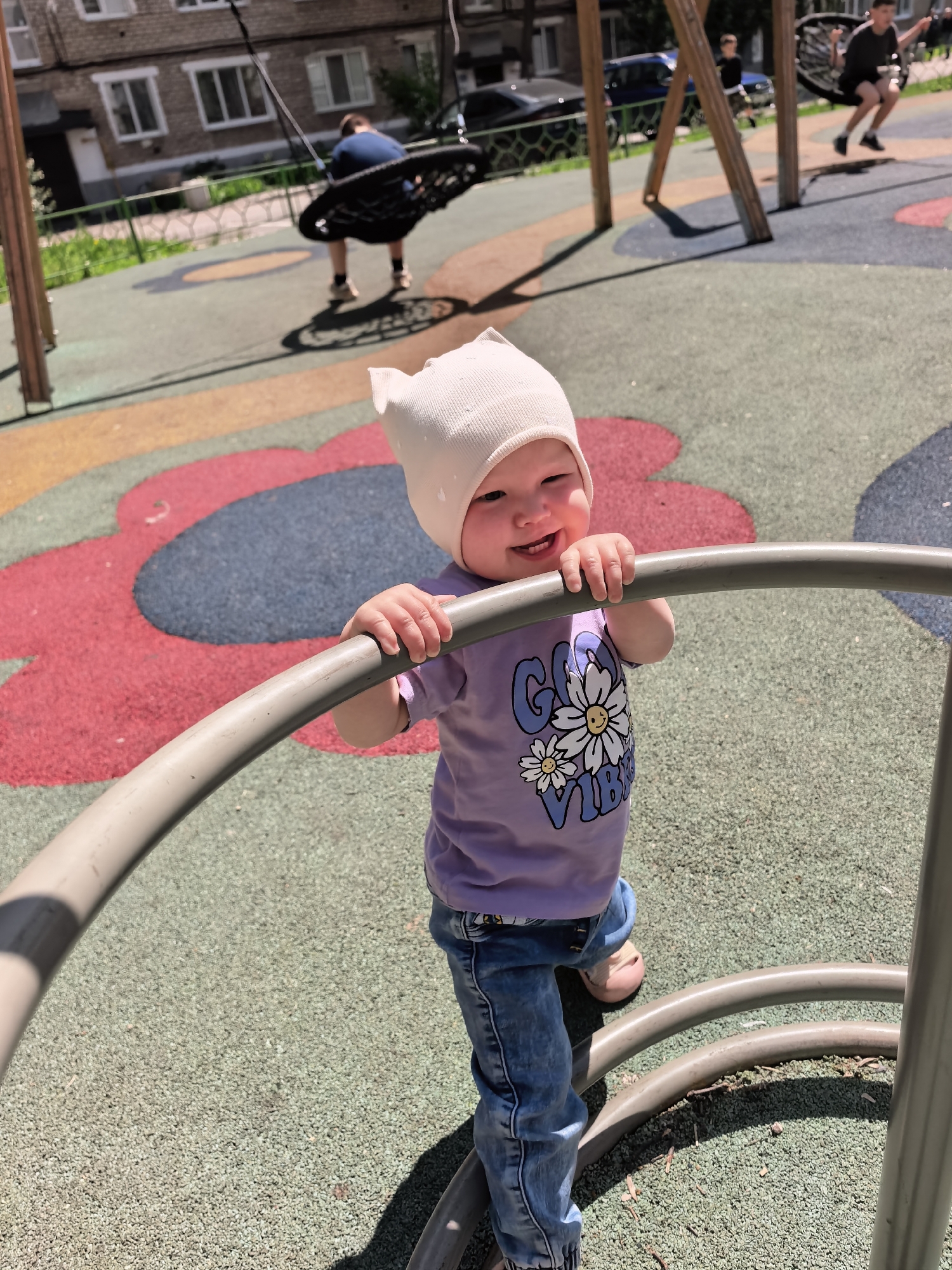 A young child wearing a white cap and a purple t-shirt with a daisy graphic stands on a playground. The child is smiling and holding onto a metal railing. The playground has a textured surface with red and blue circular patterns. In the background, other children are visible on swings. The scene appears to be a sunny day, and the overall atmosphere is playful and energetic. The location is identified as a playground in Sterlitamak, Russia.
