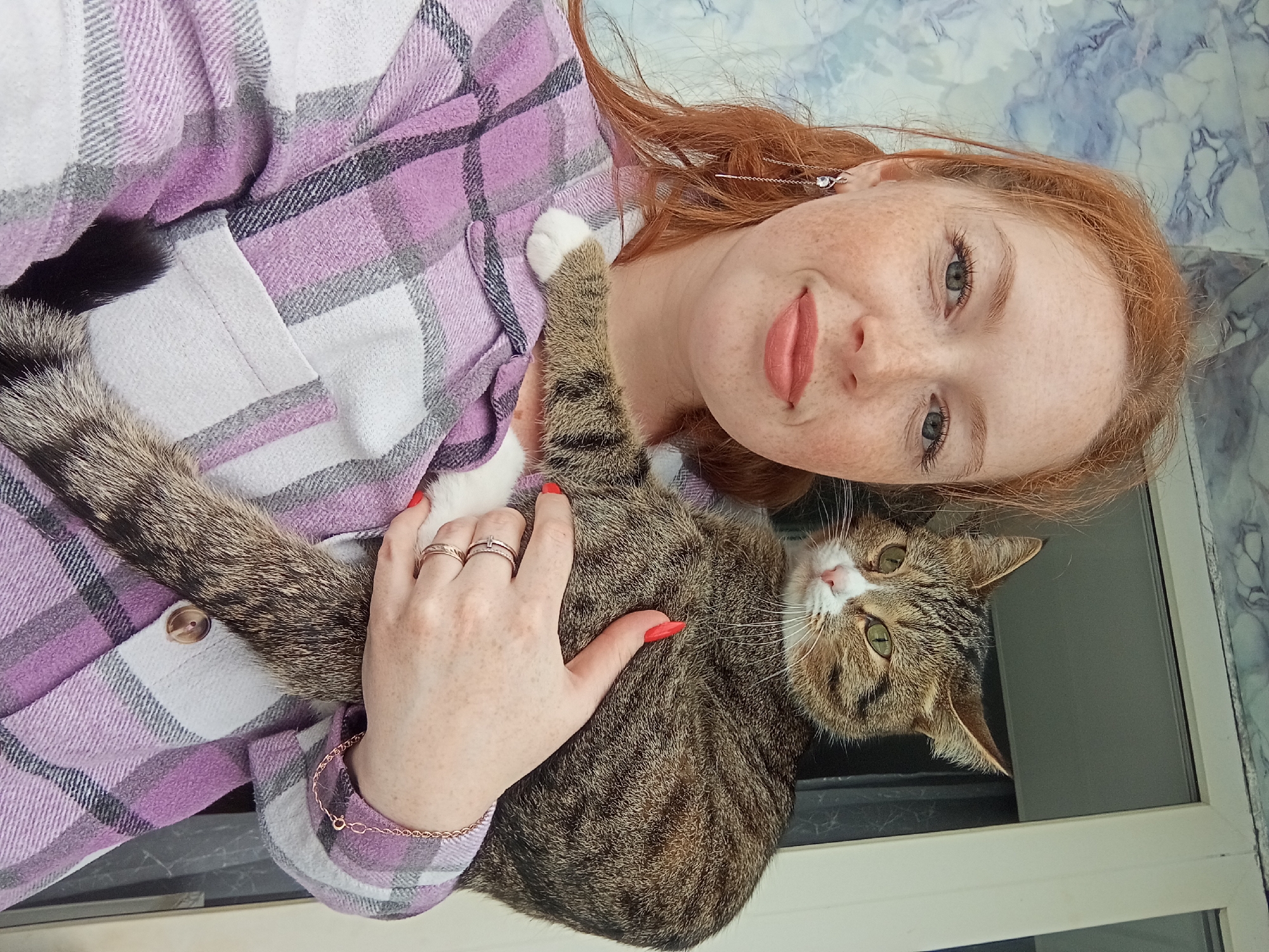 A reddish-haired woman with freckles is holding a tabby cat. She is wearing a light purple and white plaid shirt. Her hair is long and parted to the side, with long, thin, silver earrings. She has red fingernails and a rose-gold bracelet on her left wrist. Two rings are on her left ring finger. The cat is brown and gray striped with white paws. It is lying on its back and looking at the camera. The background is a mottled white and blue surface, possibly a wall or window frame. It is unknown if this photo was taken in Yurgamysh, Russia.