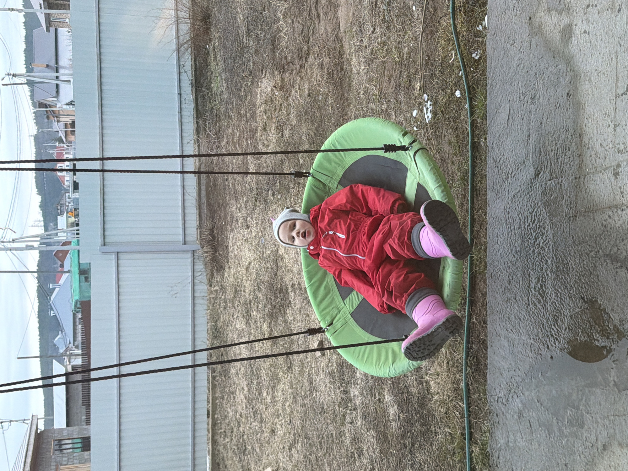 A young child is sitting in a circular, green and gray swing outdoors. The child is wearing a red snowsuit, gray hat, and pink boots. The background includes a gray fence, a grassy area, and houses in the distance. The scene is possibly located in a residential area of Chaikovsky, Russia, as indicated by the location information.