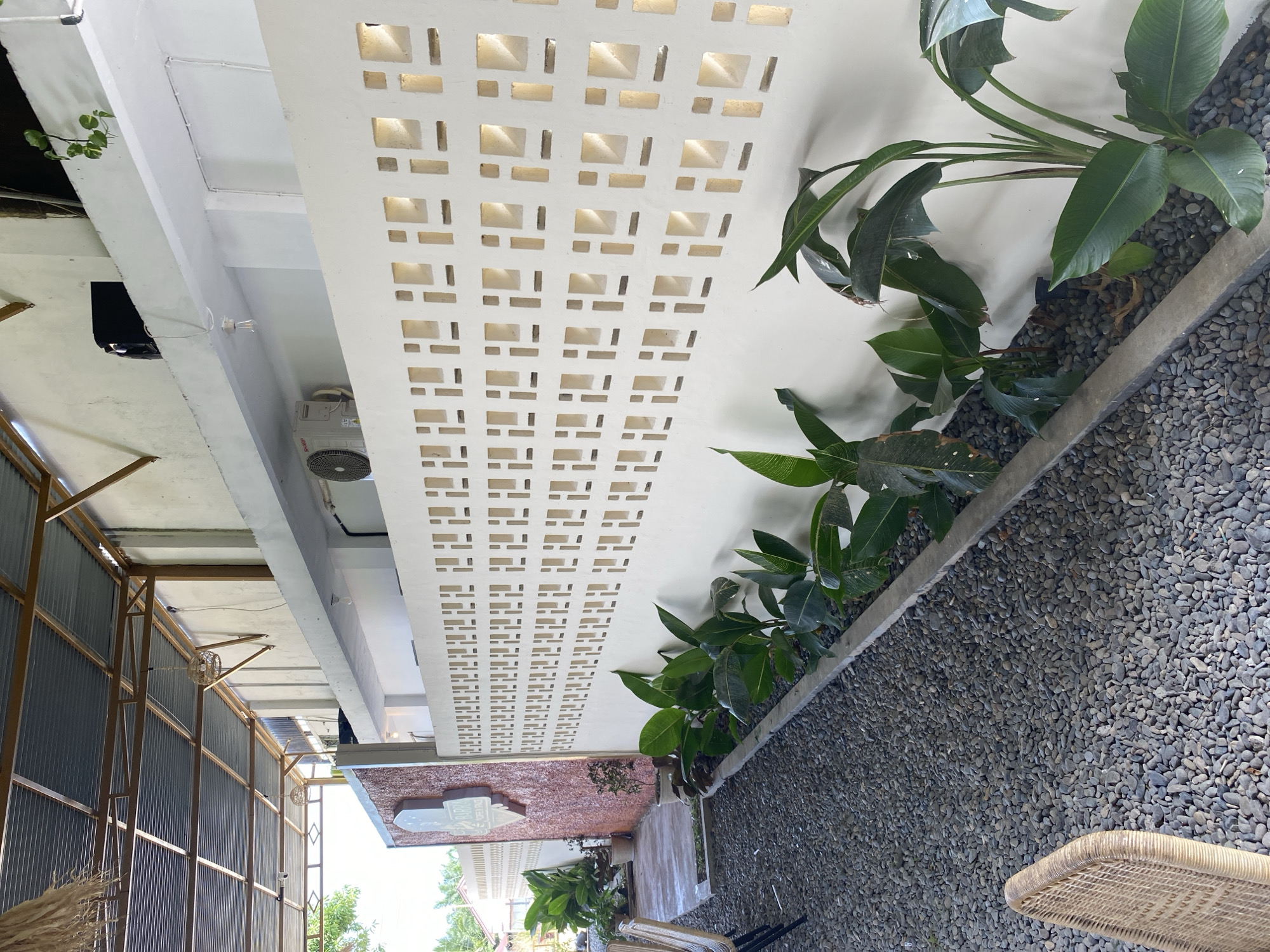 The image depicts an outdoor seating area, likely part of a cafe or restaurant, in Aluepiet, Indonesia. A white decorative wall with geometric cutouts runs along the side of the area, separating it from a gravel ground cover. Lush green plants are planted along the base of the wall. In the background, a corrugated metal roof is supported by a metal frame, suggesting an open-air structure. A sign with text, partially obscured by a wall, is visible on the left. The overall impression is one of a relaxed, tropical ambiance.