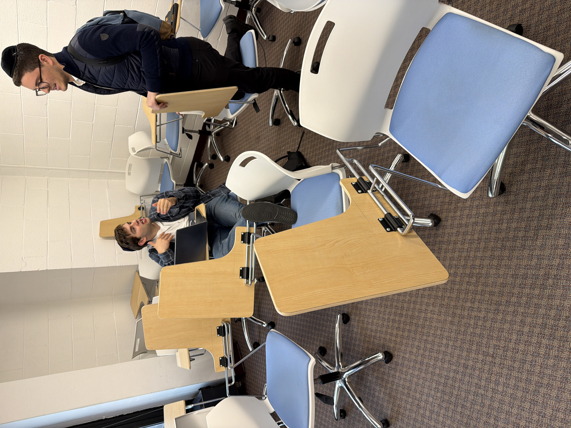 The media file captures an indoor scene, likely a classroom or study area, featuring two young men and multiple modern desks with attached chairs. The image is oriented sideways, with the subjects appearing to stand on the left wall and the floor on the right.
One young man, seen standing on the left, is wearing a dark blue vest over a dark long-sleeved shirt, dark pants, and a black yarmulke (kippah). He has glasses and a backpack slung over his shoulder. He is leaning slightly on a wooden desk, looking down and smiling.
Further into the room, another young man is seated in a chair, facing the standing man. He is wearing a plaid shirt over a white t-shirt, jeans, and a dark yarmulke. A laptop rests on his lap, and he appears to be in mid-conversation or gesturing with his hands.
The furniture consists of individual student desks, each with a light wooden top and an attached chair. The chairs have white plastic backs and light blue fabric seats, all on chrome, wheeled bases. Several empty chairs and desks are visible throughout the room, suggesting a larger learning space. The floor is covered with a brown and grey patterned carpet, and the walls are white, appearing to be made of textured blocks. The overall atmosphere suggests a casual academic or study setting. No visible text is clearly legible in the image.