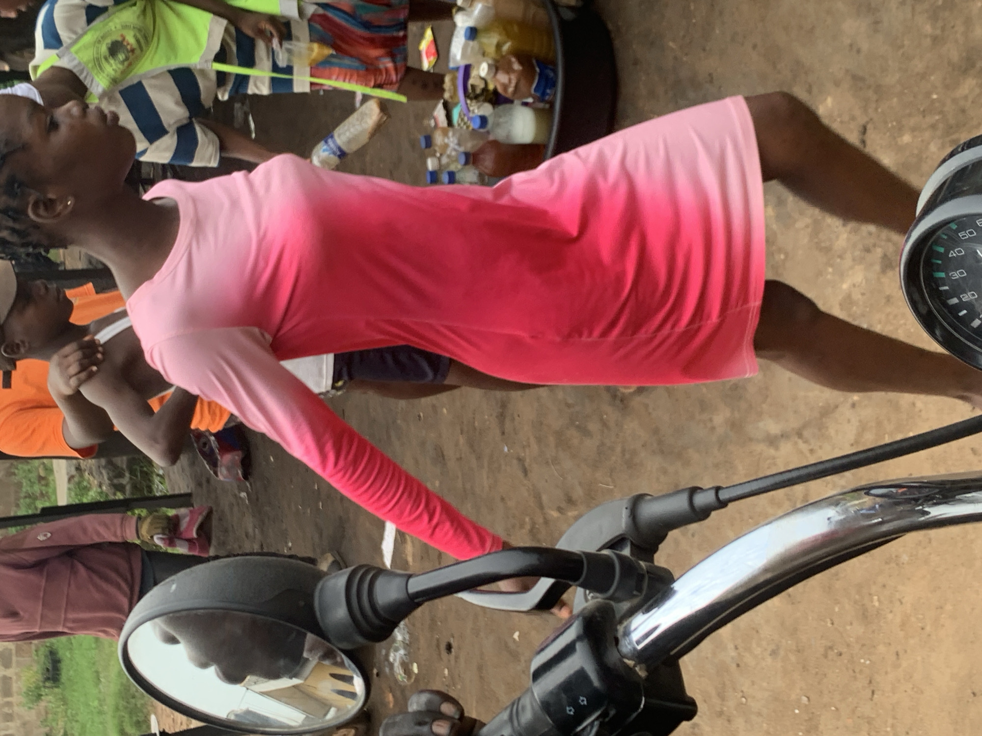 A group of people are gathered outdoors, possibly at a market, in Shagamu, Nigeria. In the foreground, the handlebars and a mirror of a motorcycle are visible. A young woman in a pink ombre dress is walking away from the camera. She has dark hair styled in braids and is wearing earrings. To her left, a person in a maroon shirt and dark pants is holding a red and white cloth. Behind the woman in the pink dress, other individuals are present, including someone wearing an orange shirt and another in a blue and white striped shirt with a neon green vest. A large basin filled with various bottles of liquids and other items is situated to the right of the woman in the pink dress. The ground appears to be dirt.