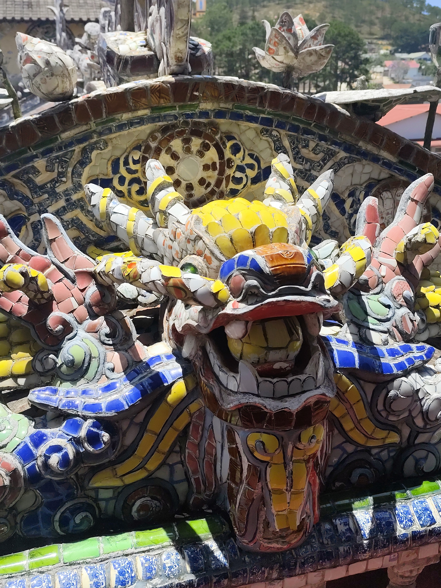 This image depicts a close-up of a colorful mosaic dragon sculpture, likely part of a larger decorative structure, in Xuân Thành, Vietnam. The dragon is composed of numerous individual mosaic tiles in yellow, blue, red, green, white, and brown hues. The dragon's head is the focal point, with an open mouth displaying teeth. The head is flanked by other dragon heads, and there is complex mosaic work in the background, potentially forming architectural elements. The entire structure appears to be outdoors.