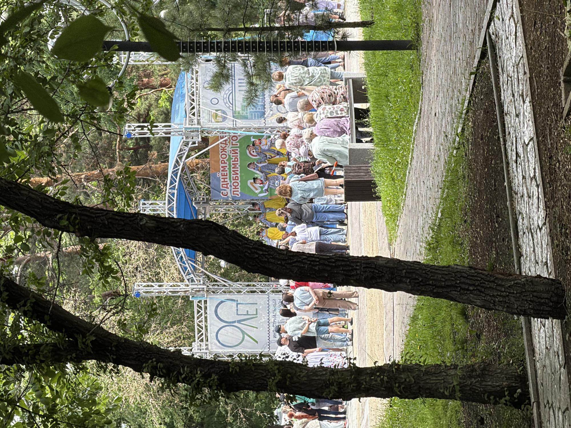 The image depicts an outdoor event in a park, likely a city celebration, with a stage set up in the background. A banner on the stage reads "90 ЛЕТ" (90 years) and "С ДНЕМ РОЖДЕНИЯ ЛЮБИМЫЙ ГОРОД" (Happy Birthday Beloved City). People are gathered in front of the stage, some watching the performance on stage, others socializing. In the foreground, a paved pathway winds through a grassy area with trees. The overall atmosphere suggests a festive community gathering. The presence of the banner celebrating the city's 90th anniversary strongly indicates this event is taking place in Khabarovsk, Russia, as it is known for celebrating such milestones.