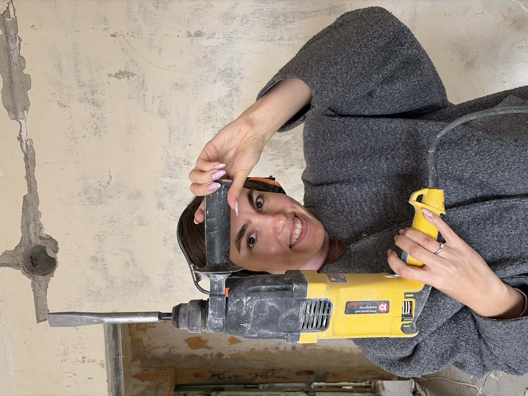 A woman stands with a yellow electric jackhammer. The jackhammer is positioned against a wall with plaster damage and holes. The woman is smiling and holding the jackhammer in both hands. She has painted fingernails and wears a ring on her left ring finger. The environment appears to be an indoor construction or renovation setting. The jackhammer has Russian writing and the text "Izhevsk". The jackhammer has the mark of the Kalibr plant, so this plant is located in the Russian city of Izhevsk.
