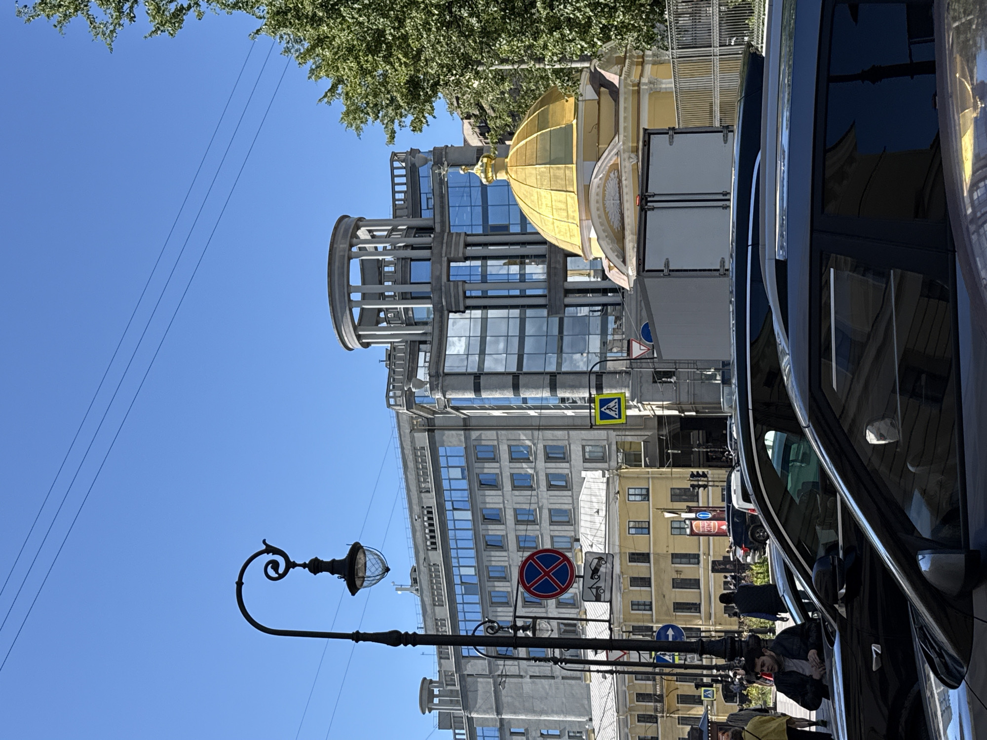 This image captures a street scene in Saint Petersburg, Russia, likely on a sunny day with clear blue skies and some tree foliage visible. The perspective is from street level, looking upwards at several buildings and a dark-colored car in the foreground.
The buildings showcase a mix of architectural styles. One prominent structure features a golden dome with a cross, suggesting a religious or historical building. Adjacent to it is a modern glass-fronted building with a circular architectural element on top. Further down the street, other buildings display classic Saint Petersburg architectural elements with repetitive window patterns.
Several traffic and street signs are visible, including a pedestrian crossing sign and a circular red and blue sign with a diagonal cross, indicating "no stopping" or "no parking." A traditional-looking street lamp with a globe is also a notable object in the scene.
People are present, though most are small in the frame and partially obscured by the car. One person is leaning into the car, and others are walking on the sidewalk. The presence of cars, including a large truck with an open back, suggests an active urban environment. The overall atmosphere appears to be one of a typical day in a bustling city. No visible text is clearly legible in the image.