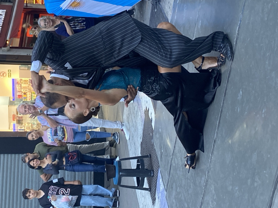 Tango dancers perform for onlookers on a city sidewalk. A man in a pinstripe suit and the woman in a teal top with a black skirt are bent dramatically during a dance routine. Behind them, a group of people is watching and a flag is overhead. The scene looks like a street performance for an audience.
