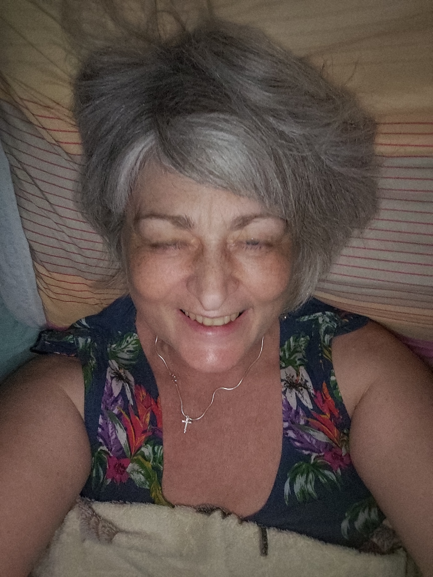 A selfie of a woman lying down, with her eyes closed and a slight smile. She has gray hair and is wearing a floral-patterned top and a silver necklace with a cross pendant. She is positioned on a bed with striped bedding. The lighting is dim and appears to be indoors. There is no visible indication of the location within the city of Krasnodar, Russia.