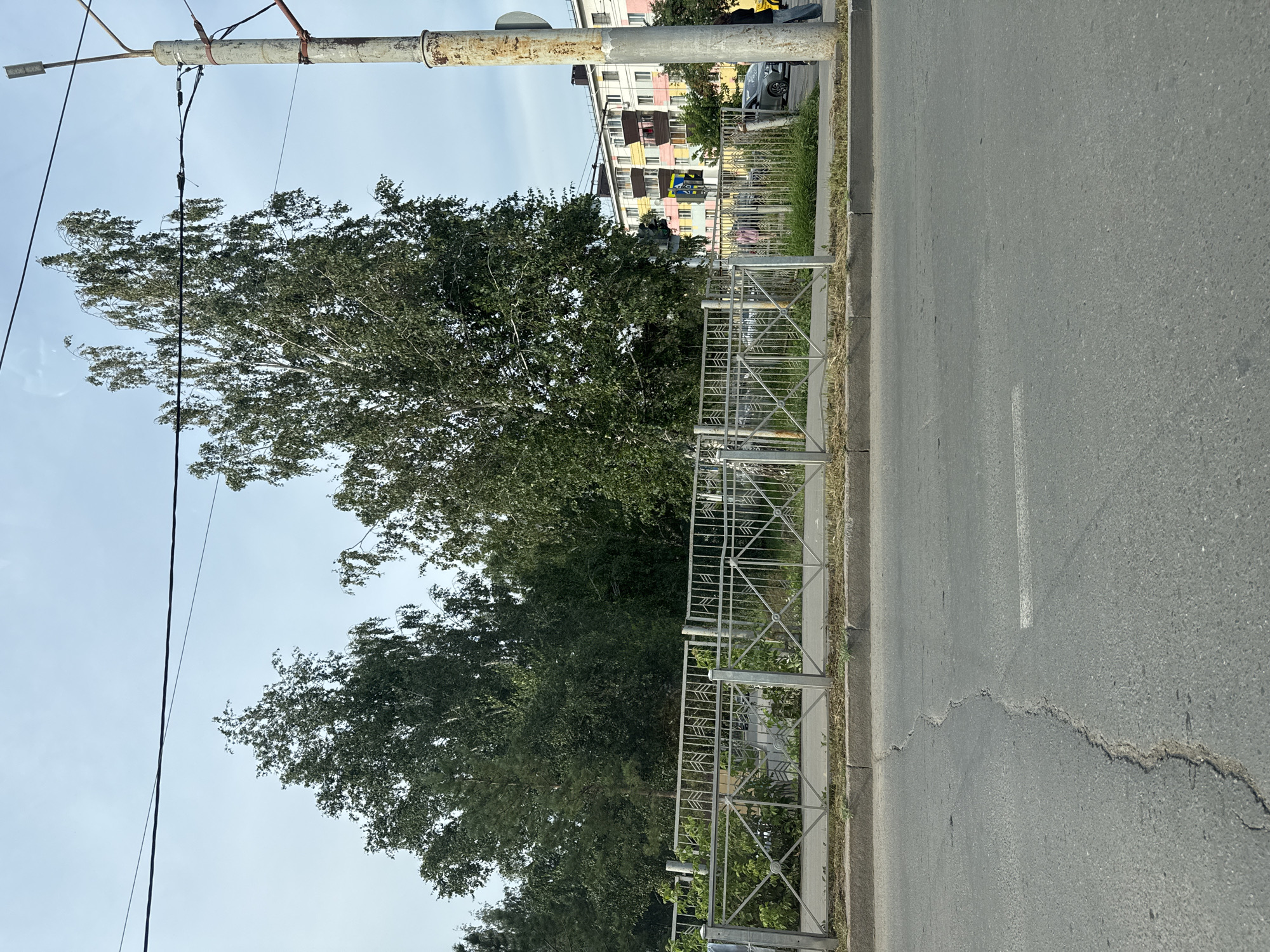 The image shows a street scene in Almetyevsk, Russia. A cracked asphalt road occupies the foreground. Behind the road, a metal fence runs horizontally across the frame, separating the sidewalk from a strip of greenery and trees. In the background, a multi-story apartment building with colorful balconies is visible, along with a utility pole. The sky is overcast.