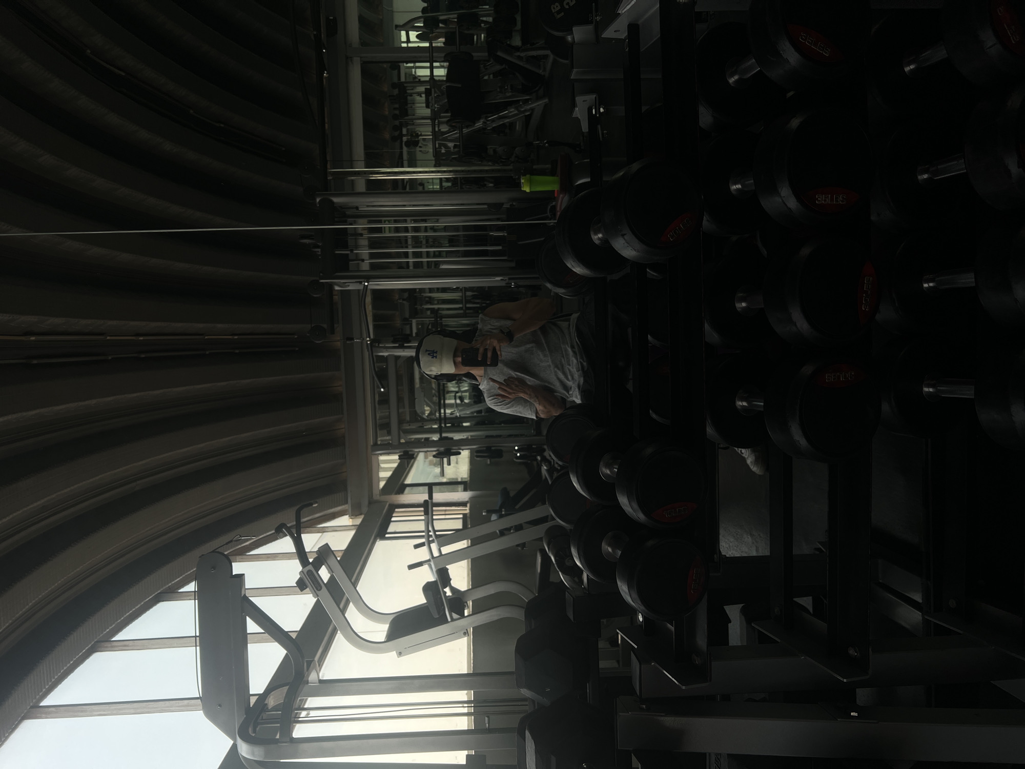 A man in a grey t-shirt and an "LA" baseball cap is taking a selfie in a gym. The gym has dumbbell racks, weight machines, and an arched ceiling with windows. The gym is located in Mexico City, Mexico.