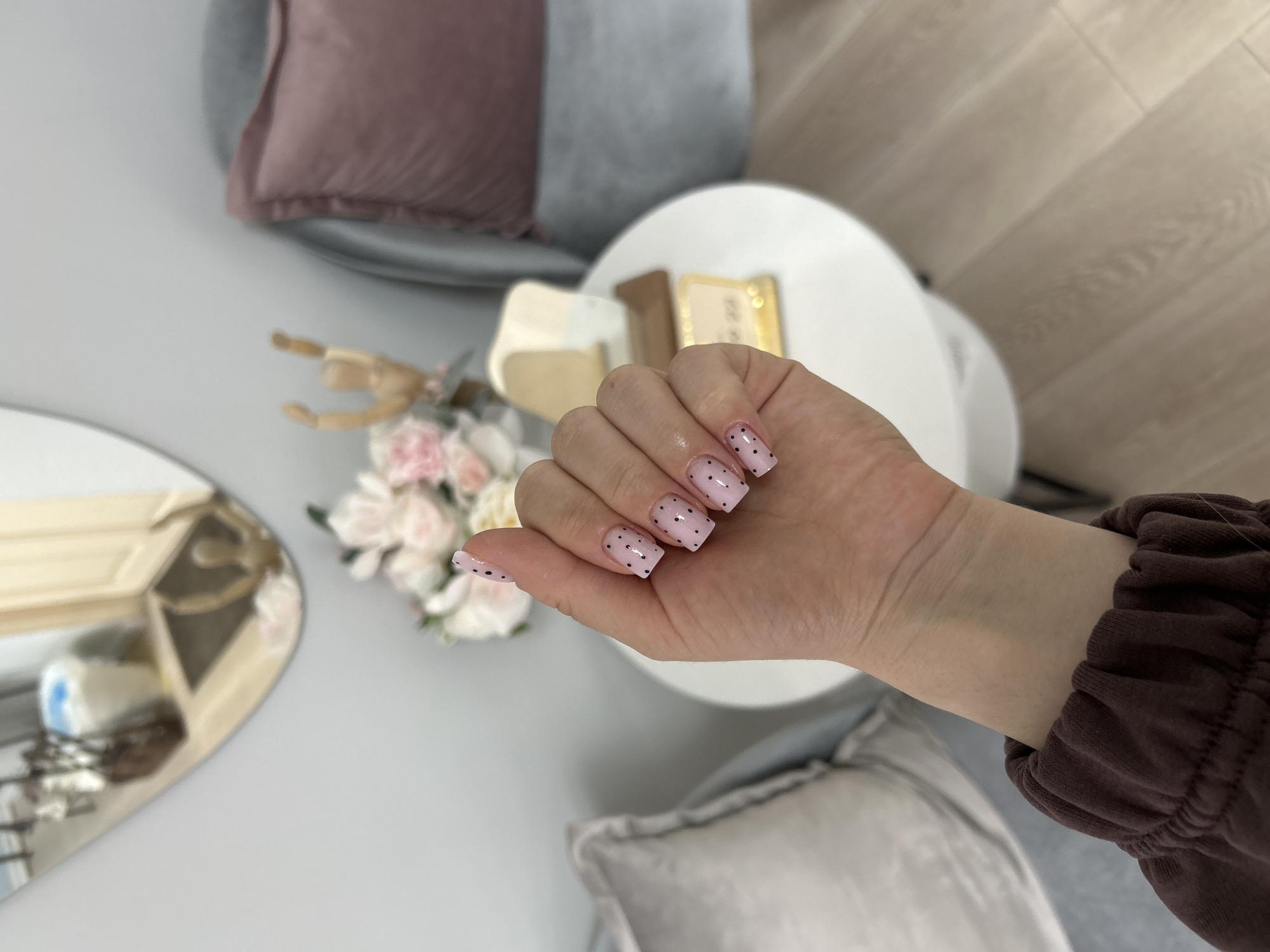 A hand is extended in the center of the frame, displaying a manicure with pale pink nails adorned with black polka dots. The hand is part of a person wearing a brown long-sleeved garment. The background includes a round white table, a bouquet of pink and white flowers, a wooden mannequin hand, and a mirror. Seating with gray and pink cushions is visible around the table. Given the location in Klin, Russia, it is likely an interior setting, such as a beauty salon.