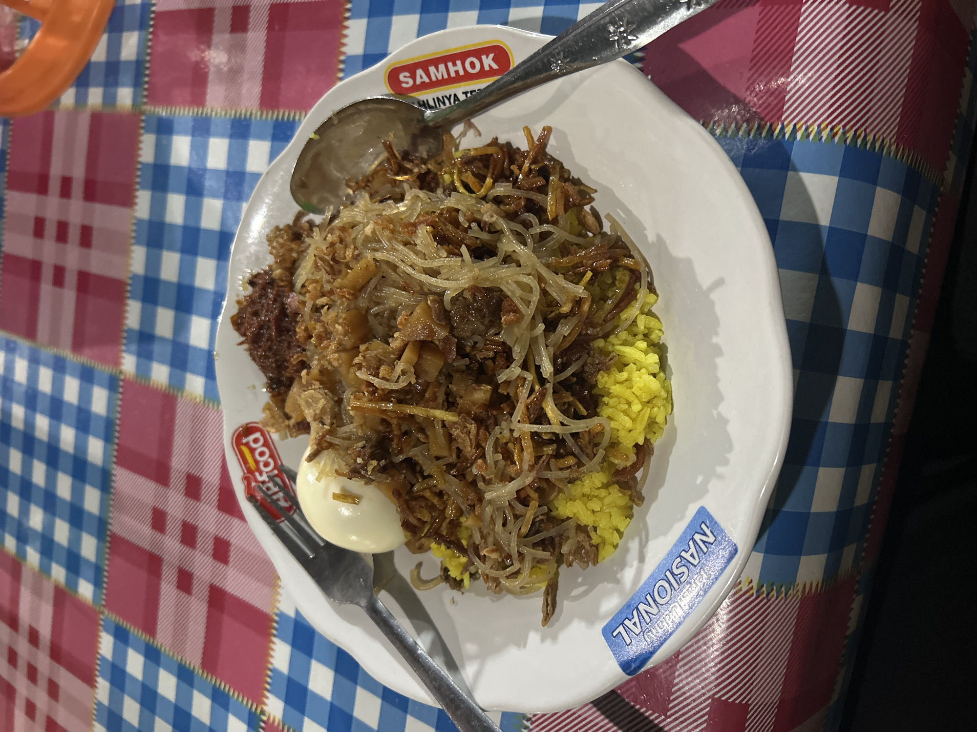 A white plate filled with Indonesian food sits on a checkered tablecloth. The food consists of yellow rice topped with noodles, shredded meat, and a hard-boiled egg. The plate has labels on it that say "NASIONAL" and "SAMHOK". A fork and spoon are placed on either side of the plate. The background is a blurry checkered tablecloth with blue, white, and pink squares. The image appears to have been taken at night, possibly at a street food stall or restaurant in Ternate, Indonesia.