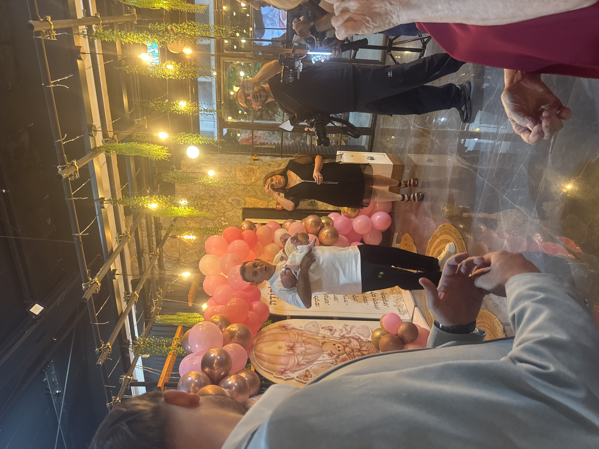 The image shows a gathering in what appears to be an indoor event space, potentially a hall or a restaurant, decorated with balloons and hanging plants. In the foreground, a man in a light-colored suit is partially visible from the back, with his hands clasped in front of him. To his right, a man in a white t-shirt is holding a baby, looking towards the camera. Behind them, a wall is adorned with a balloon arch in shades of pink and gold, and a decorative panel featuring a hot air balloon and teddy bear motif. Further to the right, a woman in a black dress stands, and a man with glasses is operating a camera on a tripod, seemingly filming the event. The environment includes stone walls, exposed ceiling structures with lighting, and a reflective floor. The scene suggests a celebration, possibly a baby naming or birthday, taking place in Migdal HaEmek, Israel.