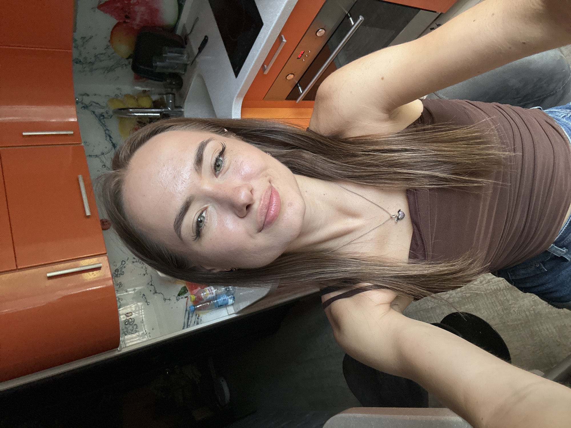A young Caucasian woman with long, brown hair is taking a selfie in a kitchen. She is wearing a brown strapless top and blue jeans, and a necklace with a heart-shaped pendant. The kitchen has orange cabinets and a white countertop with a sink. There are fruits, water bottles, and a watermelon slice on the counter. A black appliance is visible in the background. The overall lighting suggests it is daytime.