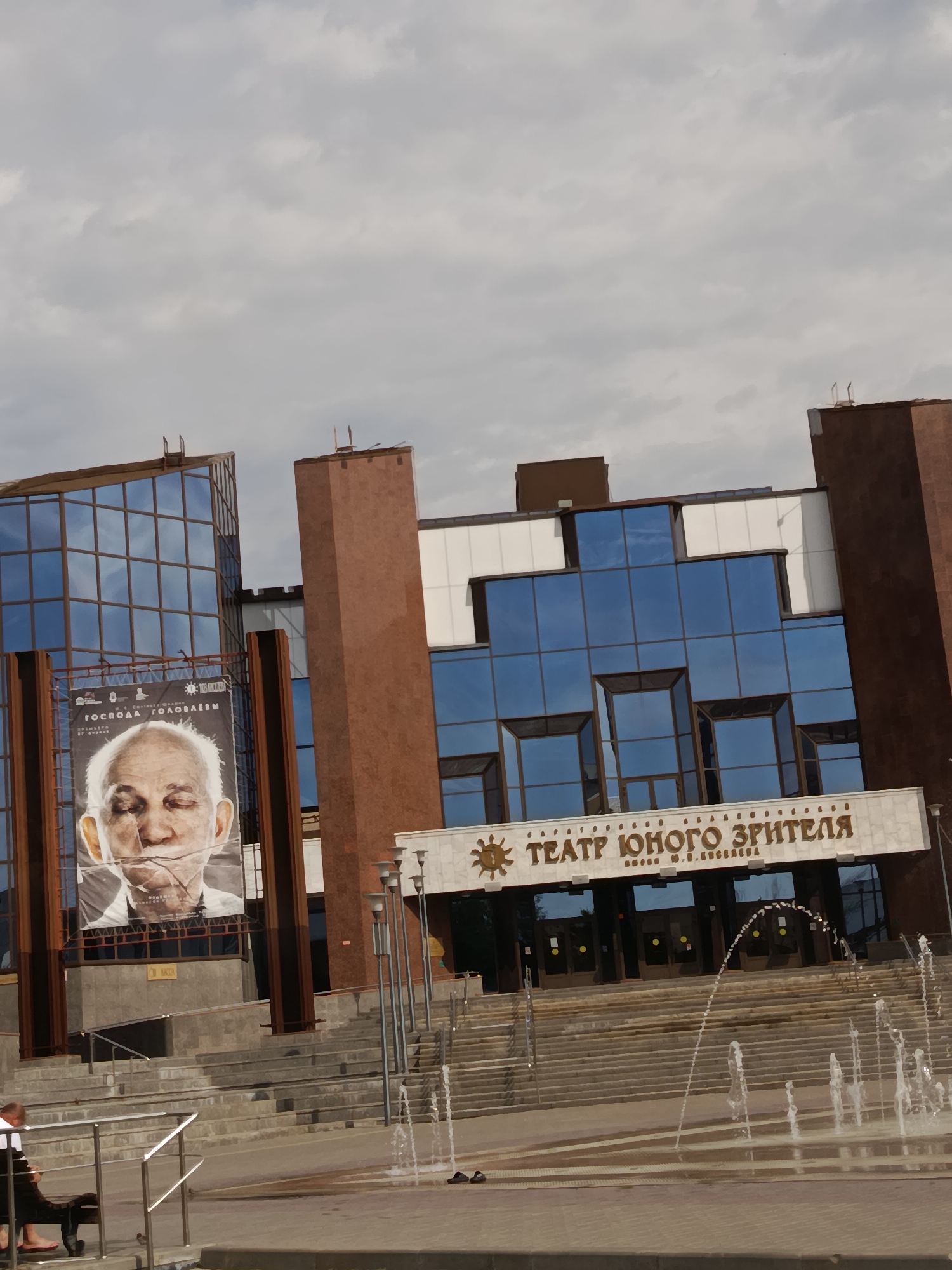 An outdoor daytime medium shot captures the facade of the Saratov State Academic Opera and Ballet Theatre in Saratov, Russia. The building features a modern glass and brown brick design. A large poster of an elderly man with his eyes closed is displayed on the left side of the building. Above the entrance, the name of the theatre, "ТЕАТР ЮНОГО ЗРИТЕЛЯ" (Theatre for Young Spectators), is prominently displayed in large, stylized gold lettering. A fountain is active in front of the building, with water spraying into the air and creating a mist. In the foreground, on the left, a person is sitting on a bench. The sky is overcast with clouds.
