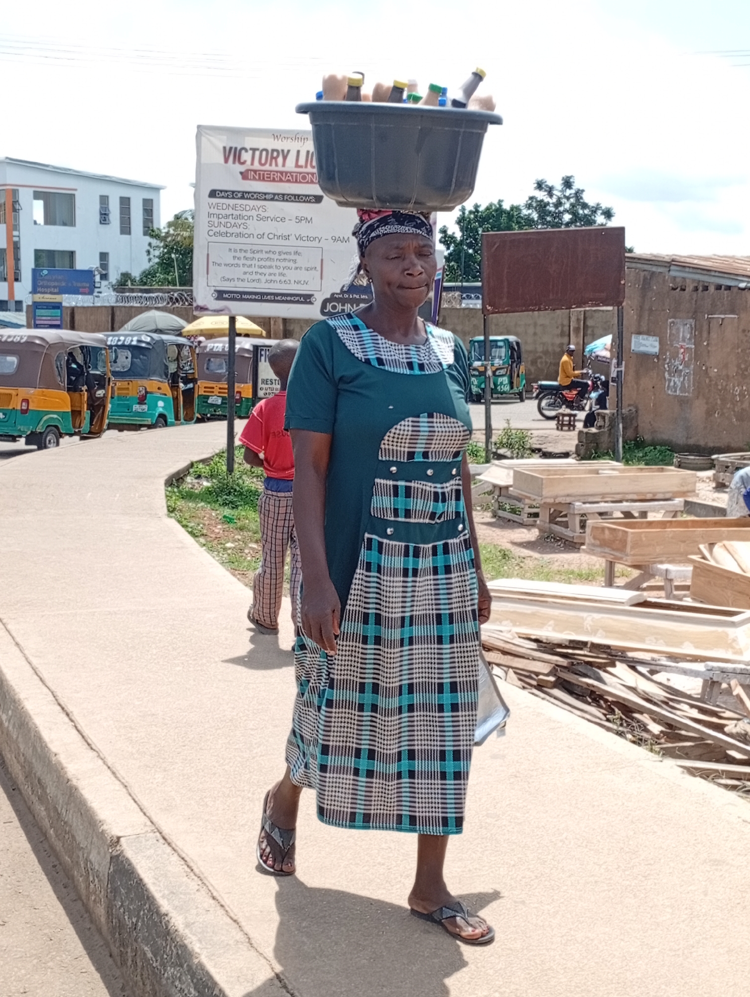 A woman is walking on a sidewalk, carrying a large basin filled with bottles on her head. She is wearing a green and black checkered dress and flip-flops. She is walking towards the viewer, her eyes are closed, and her mouth is slightly open. In the background, there are several auto-rickshaws parked along the street, and a sign that reads "Victory Light International." The environment appears to be an urban setting, possibly in a market area, with buildings and some vegetation visible. There are also wooden structures that appear to be coffins in the background. The lighting suggests it is daytime, with harsh shadows cast on the ground.