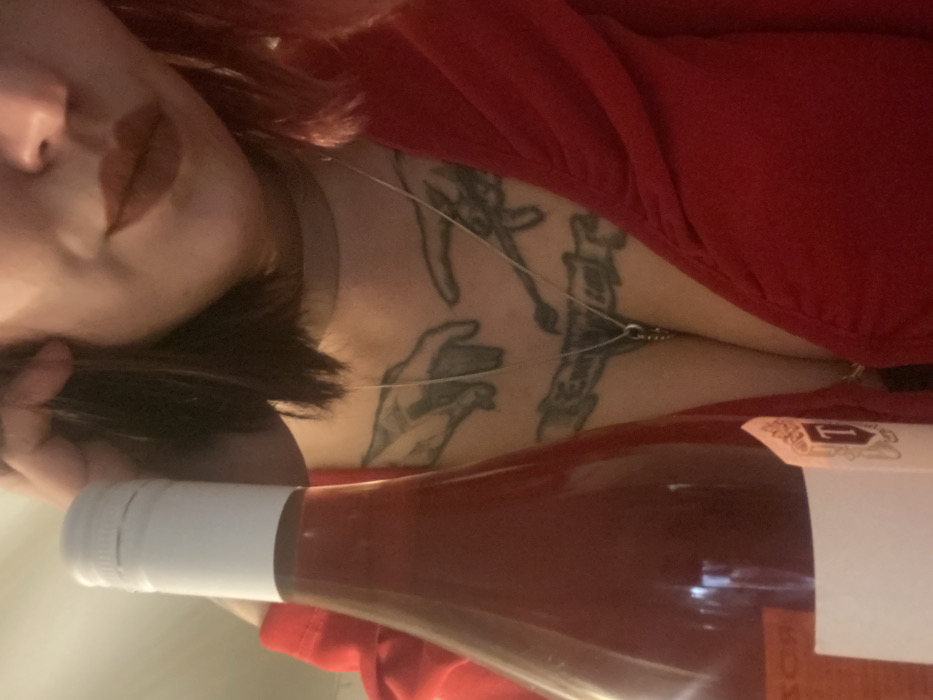 The image shows a woman in a red top lying on her side next to a bottle of rosé wine. She has short dark hair and wears a choker necklace. Visible tattoos of hands and a candlestick adorn her chest. The bottle's white cap is prominent, with the rosé liquid within showing a light pink hue. Part of the label is visible on the bottle's body. The scene is dimly lit.
