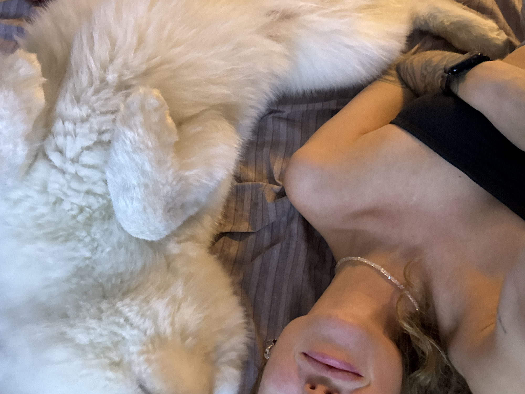 The image shows a close-up view of a woman lying in bed with a large, fluffy white dog. The woman is wearing a black strapless top and a delicate beaded necklace. Her left arm is visible, and she has a tattoo on her forearm. She is looking upwards and to her left, with her lips slightly parted. The dog's fur is white and appears very soft. The background is a striped brown and grey comforter. There are no specific indicators within the image to determine the location in the city of Chaikovsky, Russia.