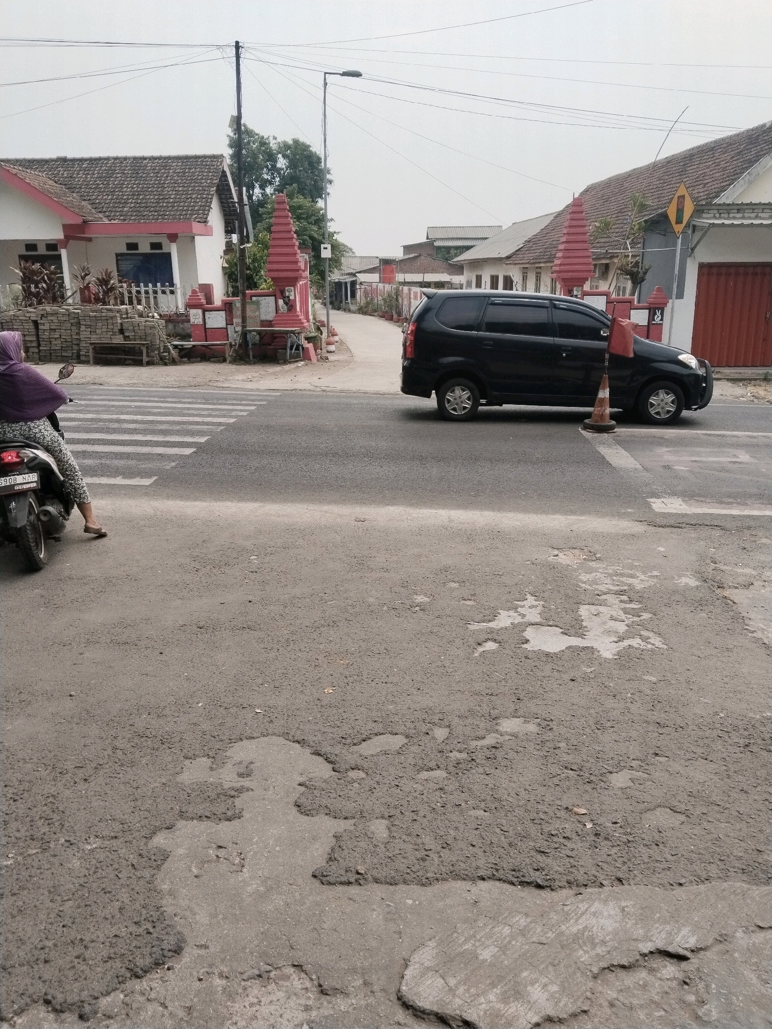 The image depicts a street scene in Pandanarum, Indonesia, characterized by weathered asphalt, residential buildings, and a zebra crossing. In the foreground, the road surface shows significant damage with numerous potholes and patches of repaired asphalt, indicating a state of disrepair. On the left, a motorcycle is parked, and a person wearing a purple headscarf and patterned leggings is seated on it, facing away from the camera. A zebra crossing is visible across the road in front of the motorcycle.
To the right of the zebra crossing, a black minivan is stopped, and a traffic cone with orange and white markings is placed on the road near it. Beyond the road, there are several residential buildings with tiled roofs. One building on the left has a white and red facade, with stacks of bricks in front. Another building on the right has a red garage door. Red decorative pillars with a layered, conical top are placed along the property lines of some houses, some adorned with small flags. Power lines and a street lamp are visible in the background against an overcast sky. The overall environment appears to be a typical suburban or semi-urban area.