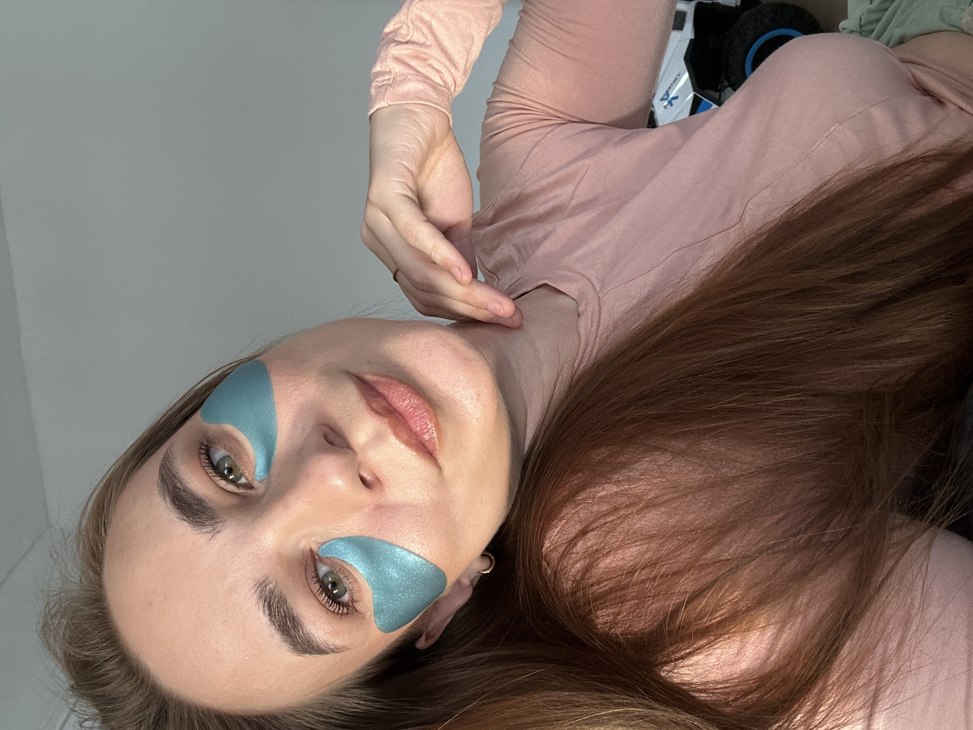 A woman is pictured indoors with a pale-pink long-sleeved shirt, applying iridescent blue triangular under-eye patches. Her long brown hair is partially visible, and she is touching her neck with her left hand. The background is light-colored and blurry. The scene's location in Kharnavy, Russia, cannot be determined from the image.