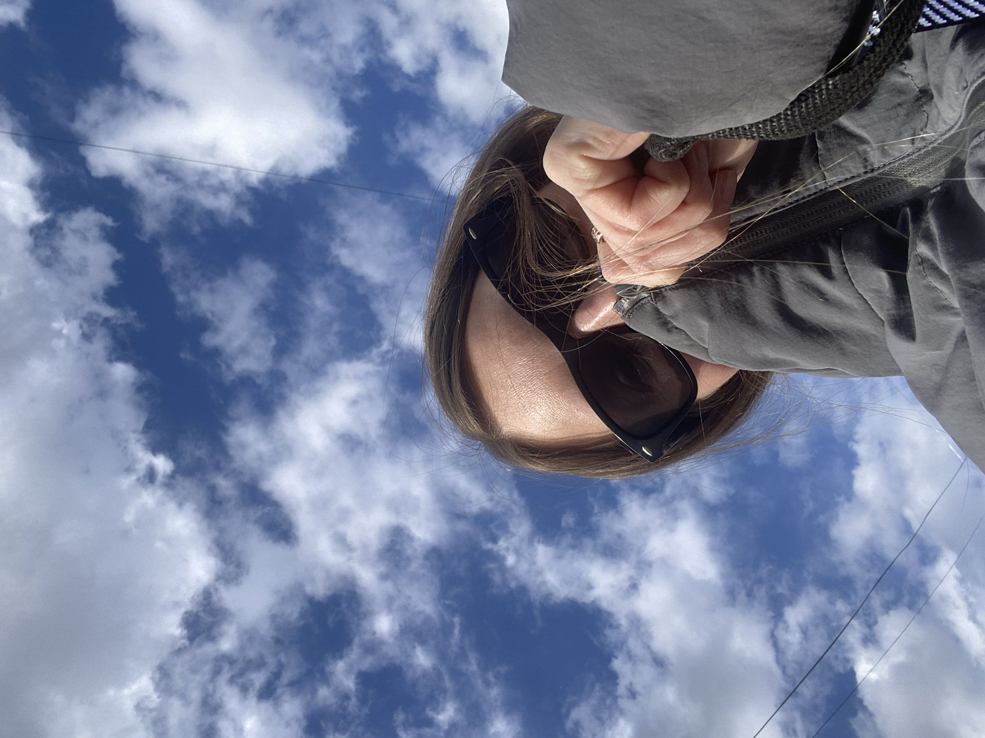 The image presents a unique, low-angle perspective, rotated counter-clockwise, capturing a person looking upwards at a daytime sky. In the foreground, a portion of an individual's head is visible, featuring long brown hair, part of their forehead, and face, largely obscured by dark black sunglasses. A hand, possibly their own, is positioned near their face, and a silver-colored ring is visible on one finger. The person is wearing a dark grey jacket or hoodie, with a zipper and a black braided strap clearly visible.
The dominant background element is a bright blue sky filled with scattered white cumulus clouds, indicative of good, sunny weather. Several thin, dark lines, likely utility wires, traverse the sky horizontally in the mid-section of the frame. The scene conveys a casual outdoor moment, possibly a selfie, taken while looking directly up. Due to the angle and sunglasses, no specific emotions are discernible. There is no visible text in the image. The provided location context is Noviy, Russia.