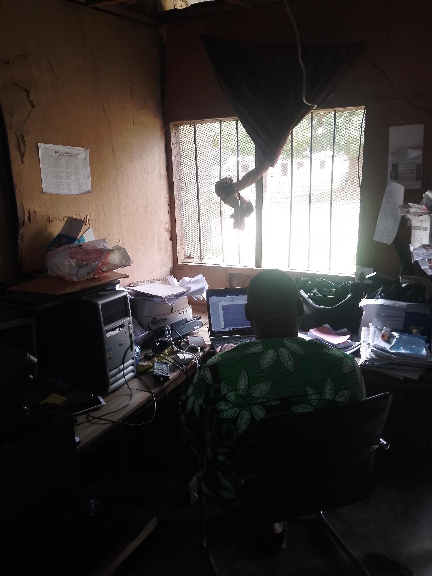 The image depicts an indoor scene, likely an office space, with a person seated at a desk in the foreground. The individual is facing away from the camera, engrossed in work on a laptop. The room is somewhat cluttered, with a desktop computer tower, stacks of papers, and various electronic accessories present on the desk. A window with metal bars is visible behind the desk, letting in a bright light that illuminates the scene. The walls appear to be made of unfinished material, suggesting a rustic or possibly under-construction environment. The overall impression is one of a functional workspace, albeit one with a degree of disarray.
