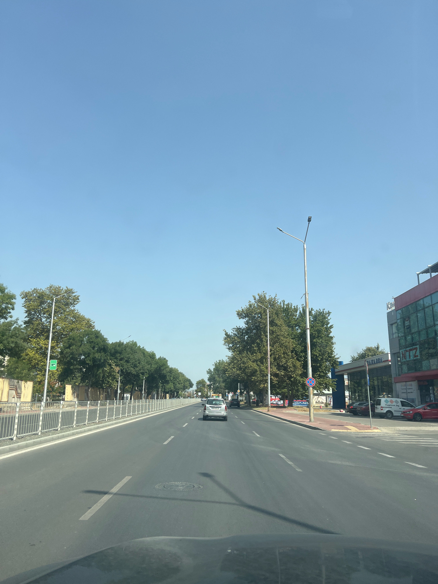 The image shows a street scene in Plovdiv, Bulgaria, on a clear day. The viewpoint is from a vehicle driving on a multi-lane road. On the left side of the road, there is a sidewalk with a metal fence and trees lining the street. Beyond the fence, there are buildings. On the right side of the road, there is a gas station with a multi-story building adjacent to it. The building on the right has a modern design with glass windows and a prominent sign that reads "NTZ". Several cars are visible on the road and parked at the gas station. The sky is blue and clear.