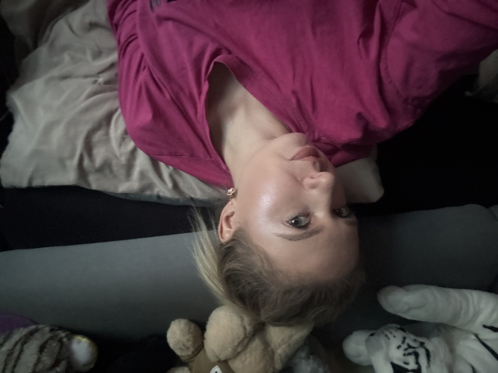 A close-up shot shows a woman from the chest up, lying down and looking at the camera. She has blonde hair pulled back in a ponytail and is wearing a pink long-sleeved shirt. A delicate gold earring with a flower design is visible on her right ear. Her skin appears smooth and somewhat luminous, possibly due to makeup or lighting. In the background, to the left, a portion of a white plush tiger toy is visible, along with a light brown teddy bear. The woman is lying on a surface covered by a beige or light gray fabric, possibly a blanket or sheet. The overall lighting is dim, suggesting an indoor setting, possibly a bedroom. The scene is set in an unknown location within an unknown city.