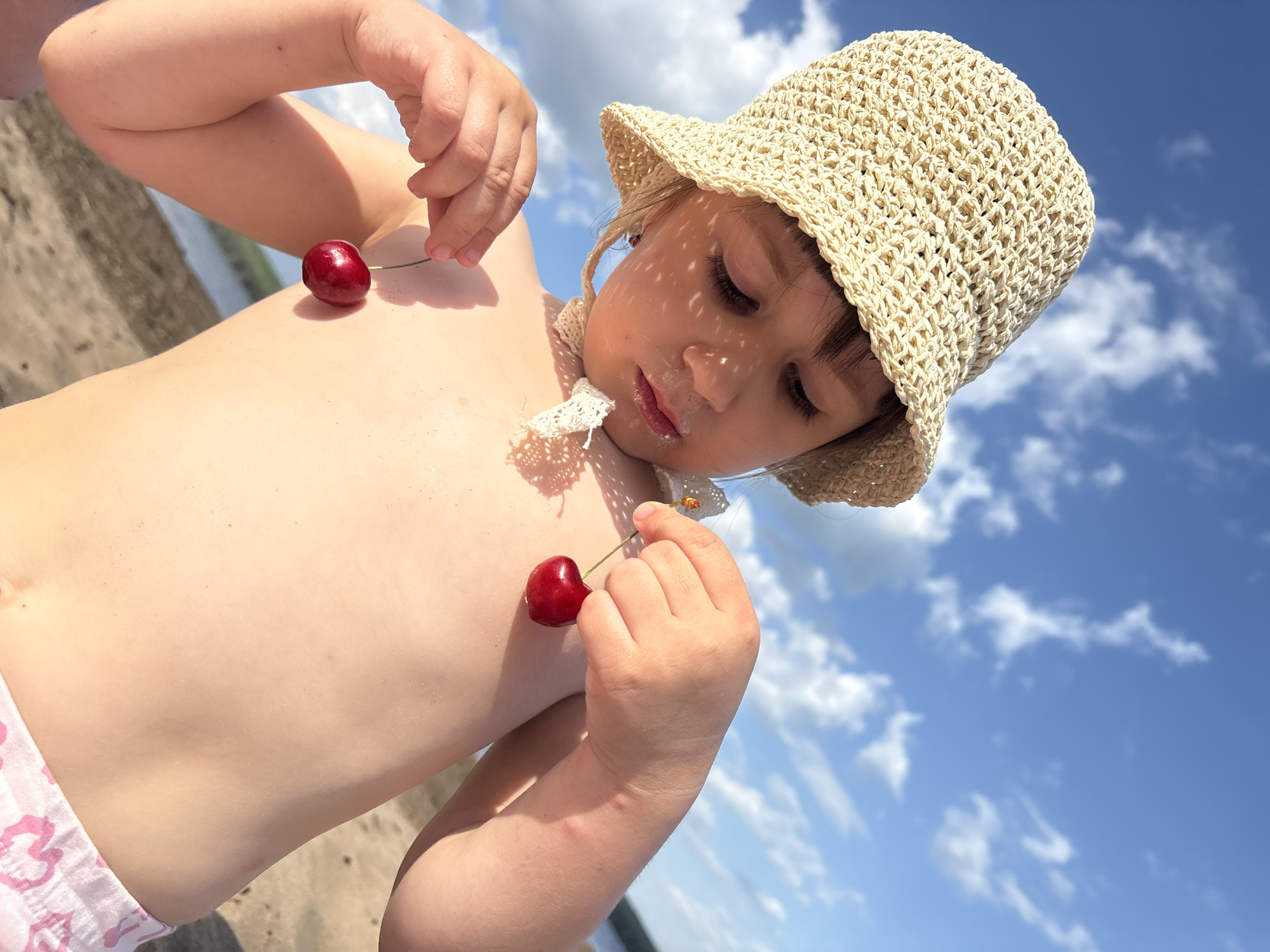 A young child lies in a partially reclined position on a beach, with a background of a blue sky dotted with white clouds. The child is wearing a woven beige hat, which casts shadows across the face. The child has pale skin and is partially unclothed, with only light pink patterned swimwear bottoms visible. Two red cherries are balanced on the child's chest, each with a green stem. The child is holding the stem of one cherry with one hand and is in motion of eating the other. The location is the city of Chaikovsky, Russia.