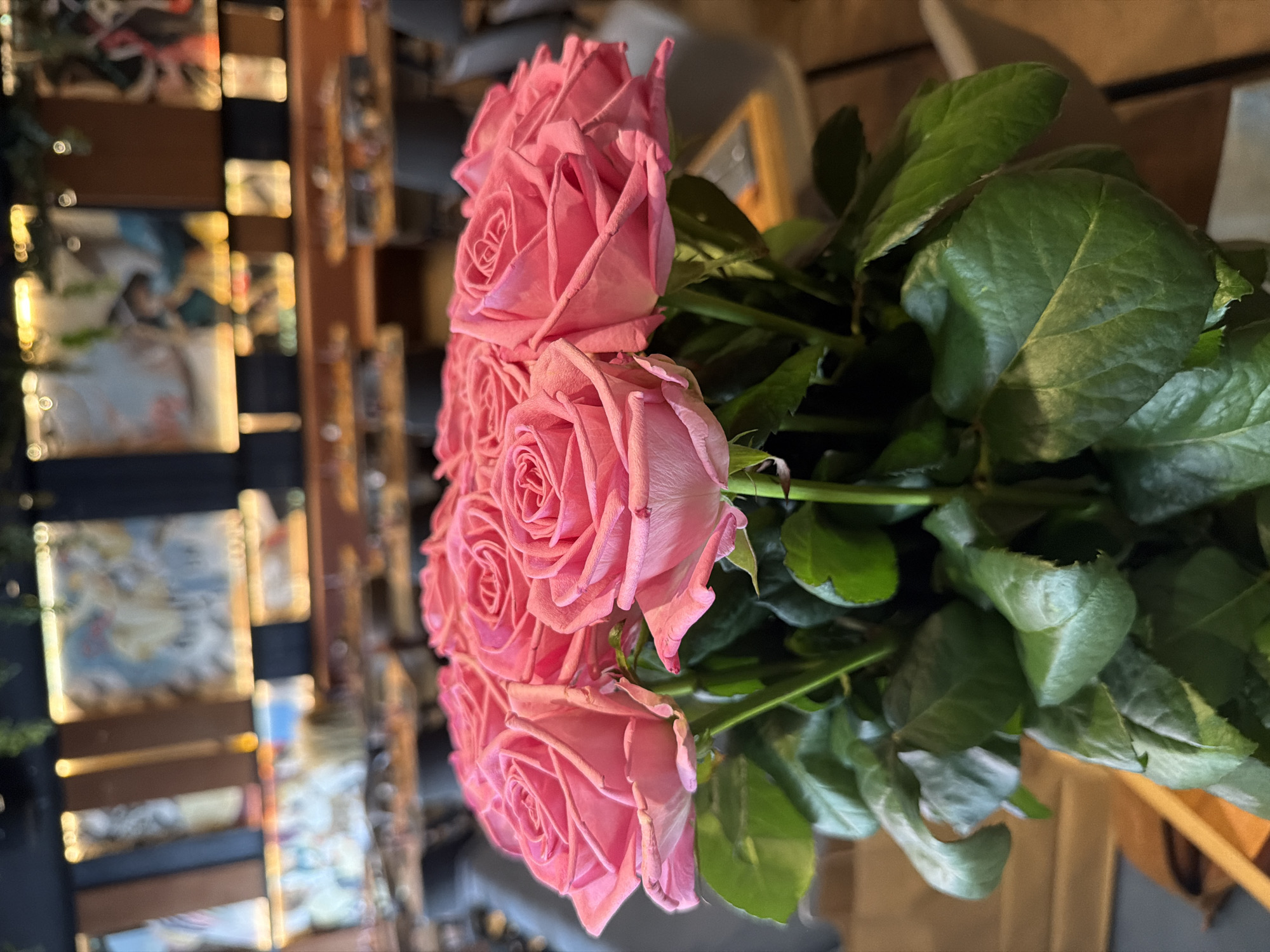 A close-up, slightly angled shot depicts a bouquet of pink roses with vibrant green leaves. The roses are in various stages of bloom. The background features a blurred view of what appears to be a multi-level display of framed pictures or posters. The location cannot be determined specifically as Tyumen, Russia with certainty.
