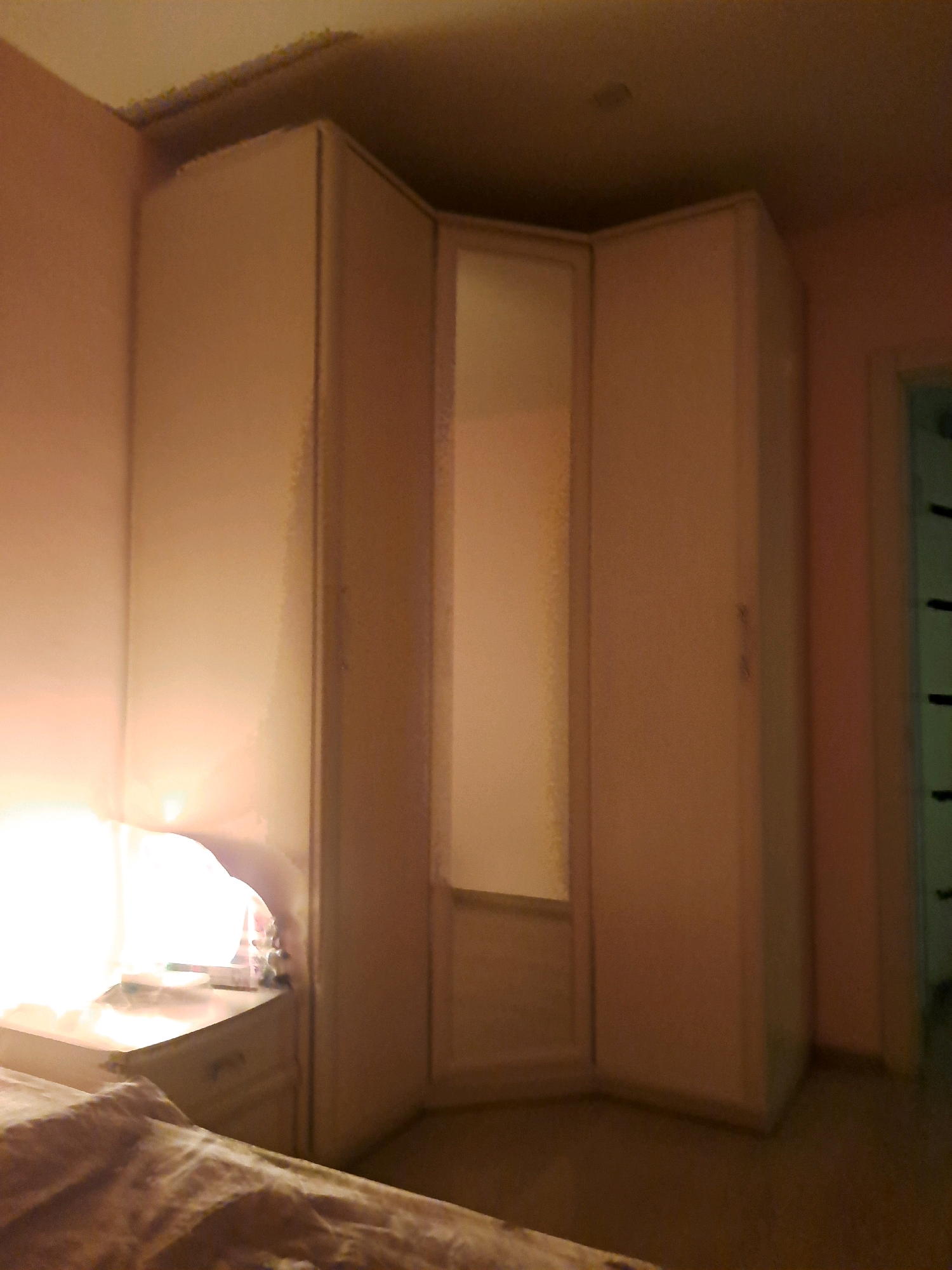 The image depicts an indoor scene of a bedroom, possibly in Irkutsk, Russia, with a focus on a large, corner wardrobe. The wardrobe is light-colored and appears to have a mirrored section in the center. To the left of the wardrobe, a bedside table is visible with a bright lamp casting a warm glow on the wall and the edge of a bed covered with a patterned blanket. The walls are painted in a pale pink or peachy hue. The floor is carpeted or has a similar soft covering. The overall lighting is dim, with the lamp being the primary light source. The scene appears to be a domestic setting, likely a residential room.