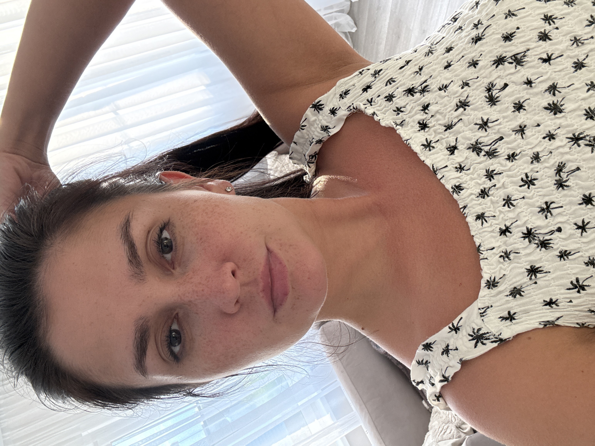 A young woman is taking a selfie. She is wearing a white top with a black palm tree print. Her hair is dark brown and pulled back. She is indoors, with bright sunlight coming through sheer curtains in the background. The woman has freckles on her face. The location in the city of Нижнеяркеево, Russia is not specified in the image.
