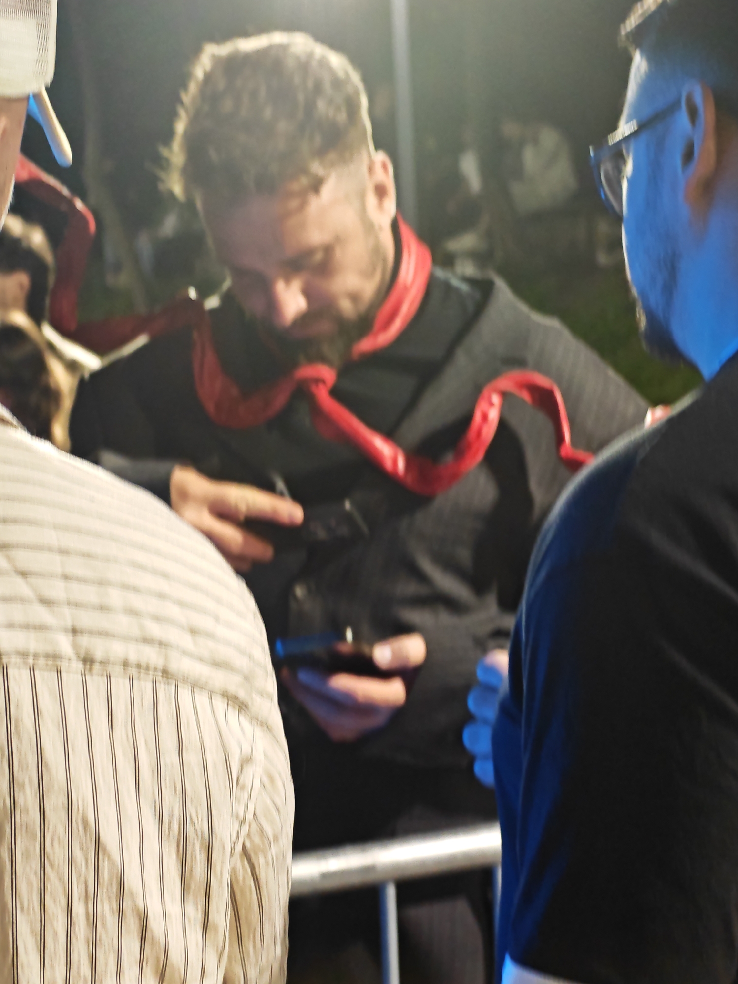 Three individuals are partially visible in the frame, with the central figure in focus. He is a man with short, textured hair and a beard, wearing a black garment with red accents around the collar and shoulders. He is holding a dark rectangular object, possibly a phone, in his right hand, with his left hand resting on his chest. To his left, another person is seen from the back, wearing a light-colored, vertically striped shirt. To the right, a third person is viewed from the side and slightly from behind, wearing glasses and a black top. They appear to be standing outdoors at night, with a blurred background suggesting trees and artificial lighting. A metal railing is visible in the foreground. The specific location in Sofia, Bulgaria, cannot be determined from the provided image.