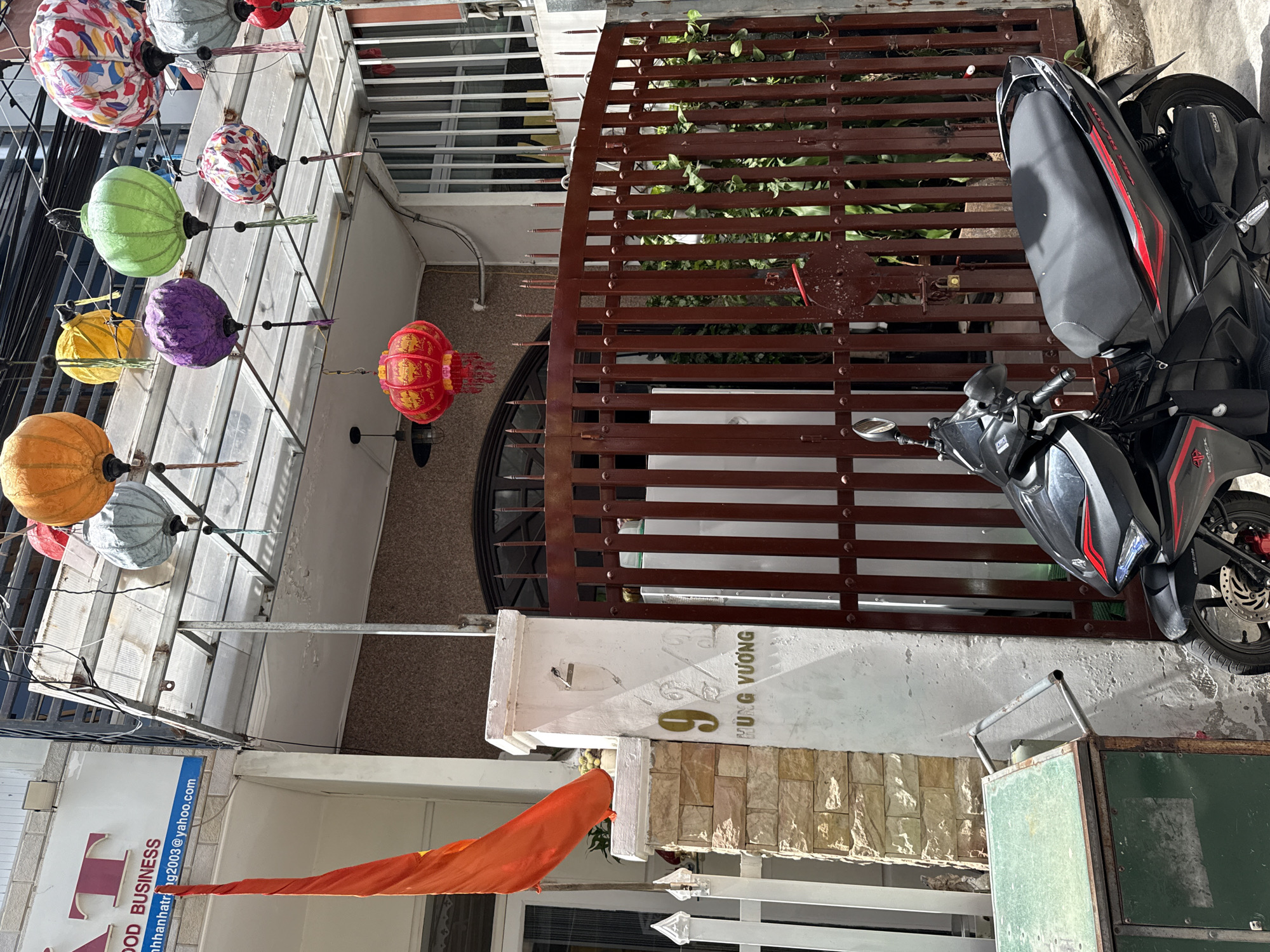 The scene depicts a street in Nha Trang Ward, Vietnam. Colorful paper lanterns adorn the exterior of a building, casting a festive ambiance. The building is adjacent to a gated property with the address "69 HUNG VUONG" displayed on the gatepost. A black motorcycle is parked behind the gate. An orange flag hangs from a building.