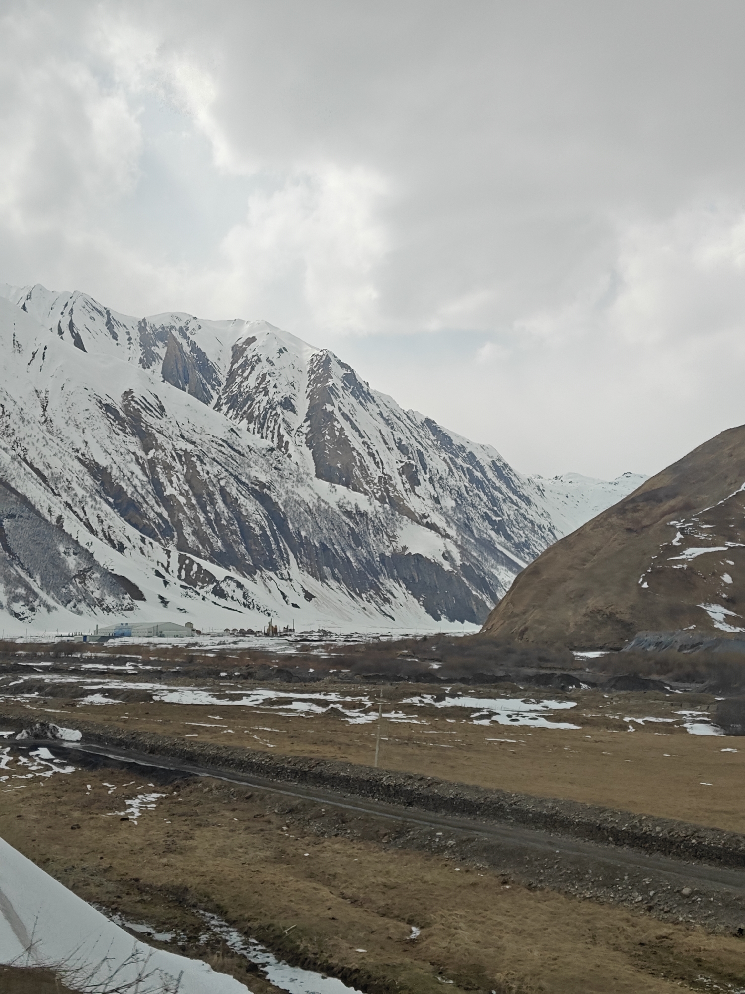 The image depicts a mountainous landscape in an unknown location. The foreground features a field of brown grass with patches of snow, intersected by a gravel road. In the middle distance, there are small structures and buildings, possibly a small settlement or industrial area. The backdrop is dominated by large, snow-covered mountains. The sky is overcast with gray clouds.