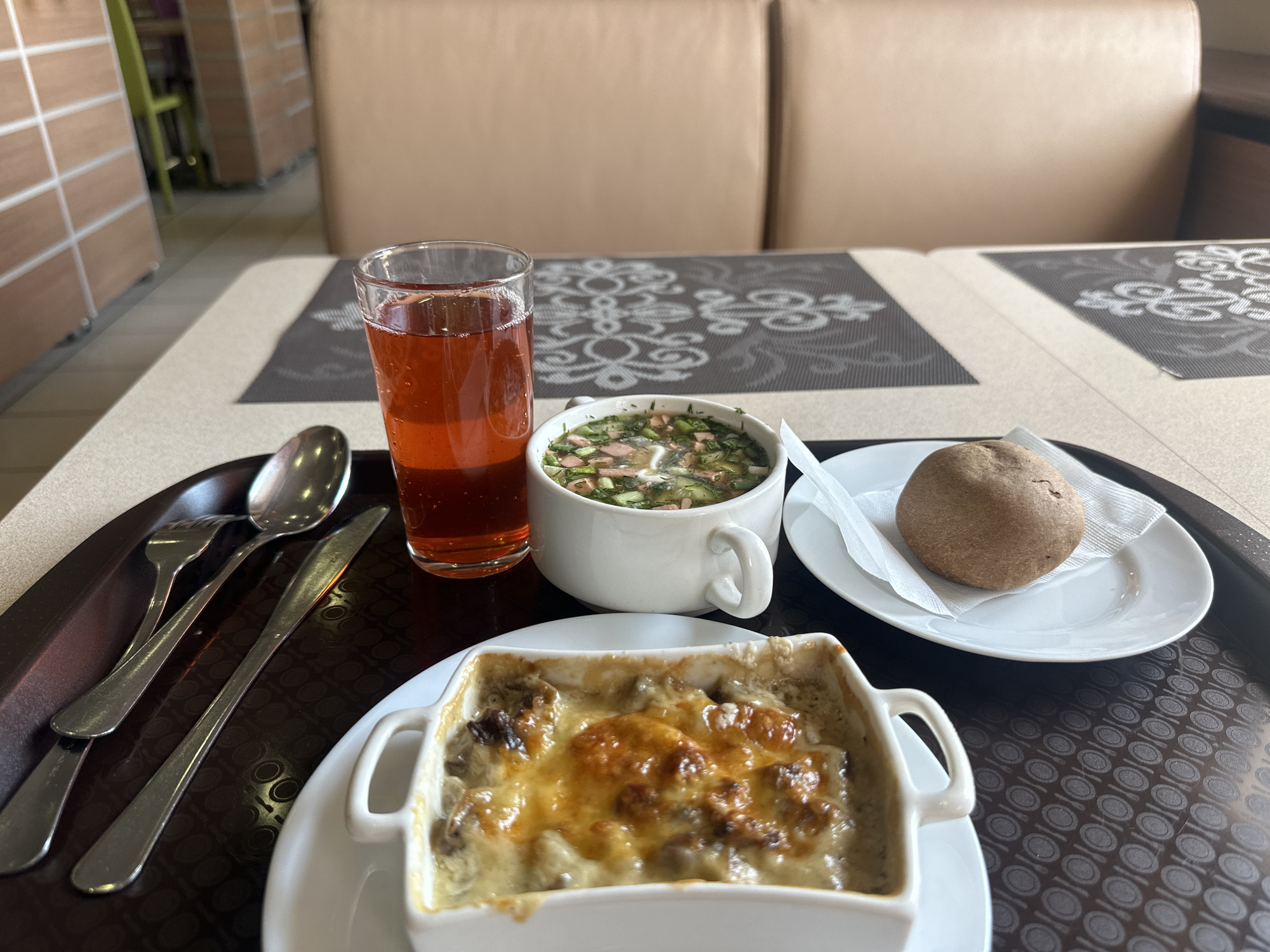 The image depicts a meal served on a brown tray on a table. The tray contains a glass of red liquid, a bowl of green and white soup, a bread roll on a small white plate with a napkin, a fork, knife, spoon, and a square white dish containing a cheesy baked dish. The table has a beige surface with a dark grey floral pattern. In the background, there is a beige couch and hints of furniture. The photograph was most likely taken in a cafeteria in Chaikovsky, Russia, given the dishes and the location.