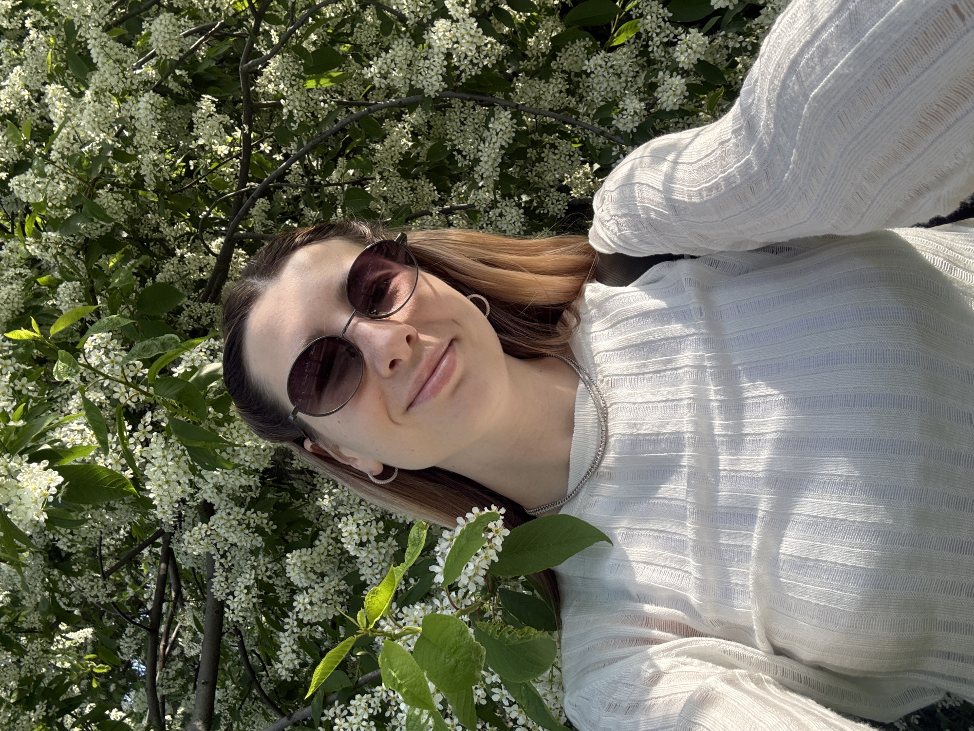 A woman wearing sunglasses and a white, textured sweater is standing amongst a tree covered in small white flowers. She is wearing hoop earrings and a silver chain necklace. She appears to be in an outdoor setting with ample natural light. The location is potentially in Moscow, Russia, though there are no clear indicators in the image confirming this.