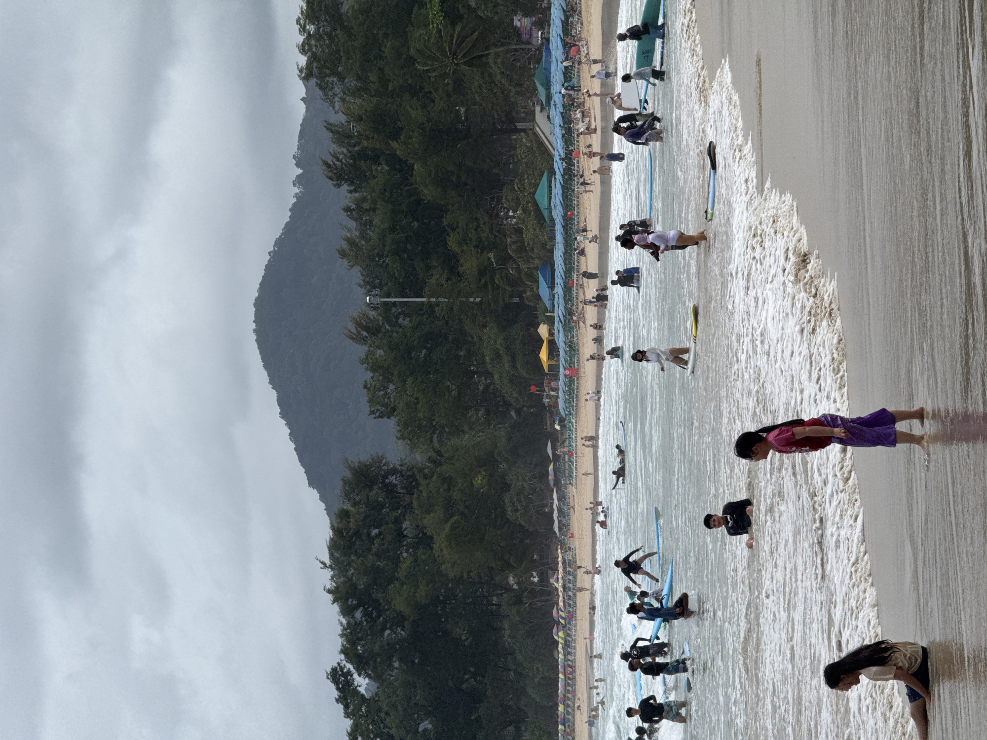 The image depicts a cloudy day at Karon Beach in Karon, Thailand. The scene is populated with numerous people engaged in various beach activities. In the foreground, two young girls are visible on the sandy shore, with one sitting and the other standing. The water is filled with a mix of individuals, some wading, others surfing. Multiple surfboards are present in the water, with several people actively paddling or positioned on them. Further out, the beach is lined with rows of colorful umbrellas and lounge chairs, indicating a busy tourist area. Behind the beach, a dense line of trees forms a backdrop, with a prominent, tree-covered mountain rising in the distance under a gray, overcast sky. The overall atmosphere is that of a lively beach day despite the subdued lighting.
