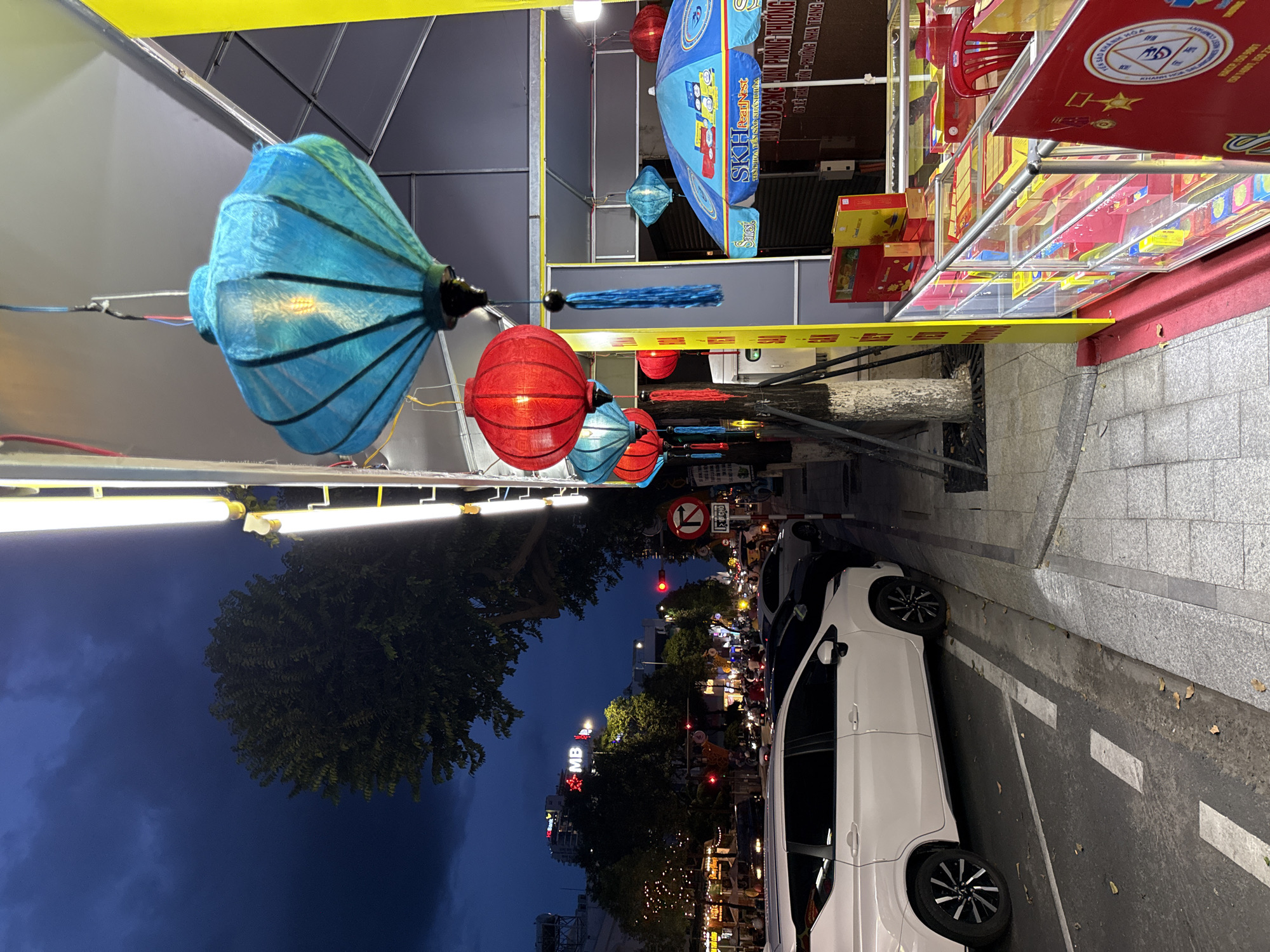 The image depicts a street scene in Nha Trang, Vietnam, at dusk. Several brightly colored lanterns, predominantly blue and red, hang from an overhead structure, illuminating the area. In the foreground, a white minivan is parked along the curb, which is marked with white lines on the asphalt. To the right, a retail stall with glass display cases showcases various boxed items, possibly food products or gifts. A red chair is visible behind the stall. The background reveals a street with traffic, buildings, trees, and streetlights, suggesting an urban environment. The sky is dark blue, indicating it is either early evening or predawn.