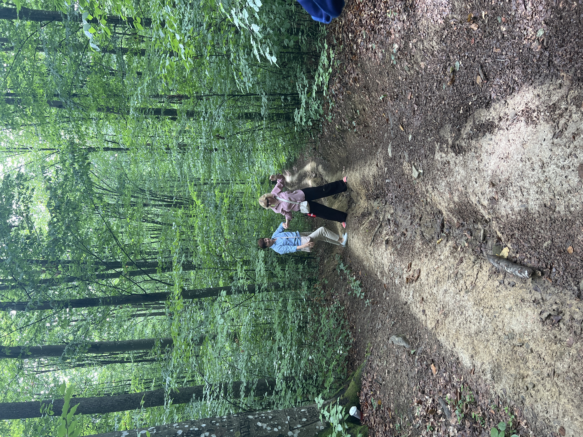 Two individuals, a male and a female, are captured standing on a dirt path in a dense forest. The male, on the left, wears a blue shirt and light-colored pants. The female, on the right, is dressed in a pink shirt and dark pants, with sunglasses perched on her head. They appear to be interacting with each other or posing for the photo. The path is uneven, with signs of mud and debris, suggesting it may be a hiking trail or a less-maintained road. The surrounding environment is characterized by numerous tall trees with lush green foliage, creating a shaded and natural setting. The image was taken outdoors, likely during the daytime, given the natural light filtering through the trees. The location is identified as the city of Pavlovo, Ukraine, implying the forest is situated within or near this area.