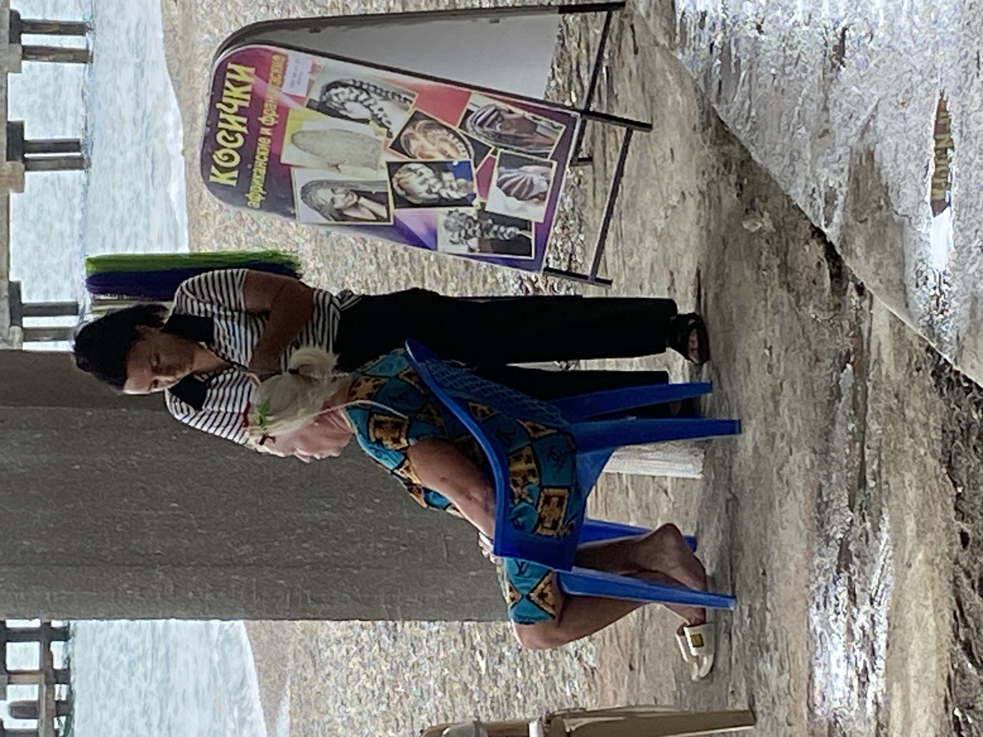 A woman is getting her hair braided by another woman on a beach in Sokhumi, Abkhazia. The woman getting her hair done is sitting in a blue plastic chair and wearing a colorful patterned dress. The woman braiding her hair is standing and wearing a striped shirt and black pants. There is a sign next to them advertising hair braiding services. The background shows the sea and a pier. The ground is concrete and wet.