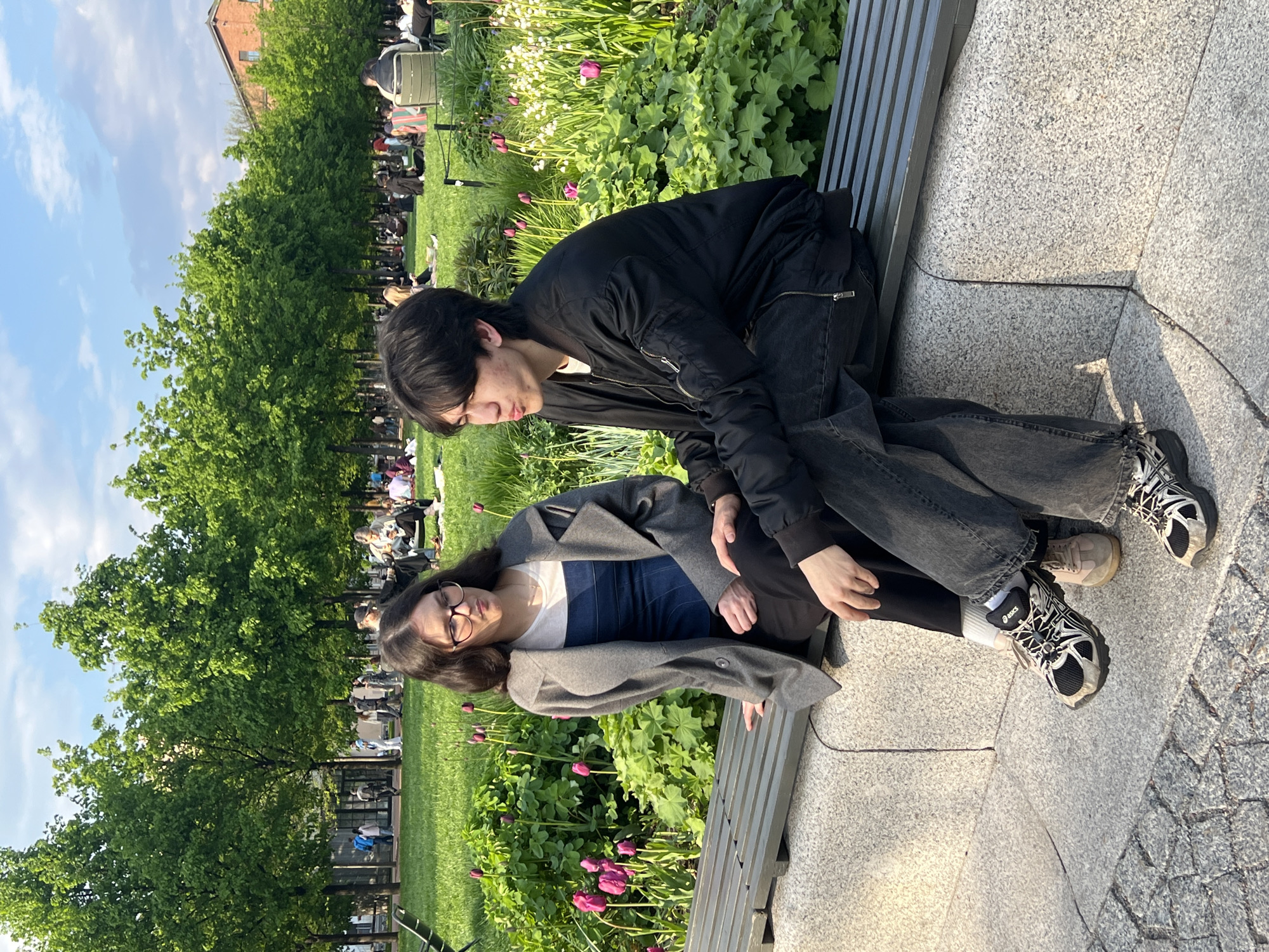 Two people are seated on a park bench in Saint Petersburg, Russia. One individual with black hair is leaning over the other, who is wearing glasses and a gray coat. The setting is a park with green trees, flowers, and other people in the background. The ground is paved with stone.