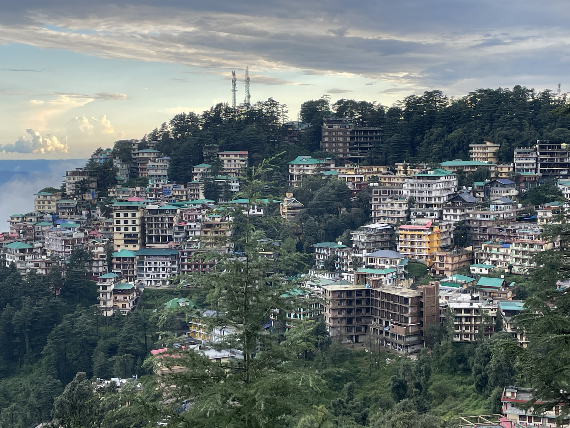 The image depicts a dense, hillside settlement in Bhagsu, India, with a vast number of multi-story buildings cascading down a steep incline. The architecture is varied, featuring structures of different heights, colors, and materials, many with flat roofs and balconies. Green-tiled roofs are a recurring element. The buildings are clustered tightly together, interspersed with lush green trees, primarily conifers, which also cover the surrounding hills.
In the background, two prominent telecommunications towers stand out against a cloudy sky. The sky itself is a mix of light and dark grey clouds, with hints of blue and illuminated patches suggesting either dawn or dusk. In the far left of the image, mist or low-lying clouds partially obscure the distant landscape, indicating a potentially mountainous or high-altitude location. There are no visible people or distinct actions taking place; the scene is a static, wide-angle view of the urban sprawl integrated with the natural environment. The overall impression is one of a densely populated town built upon a rugged, forested terrain.